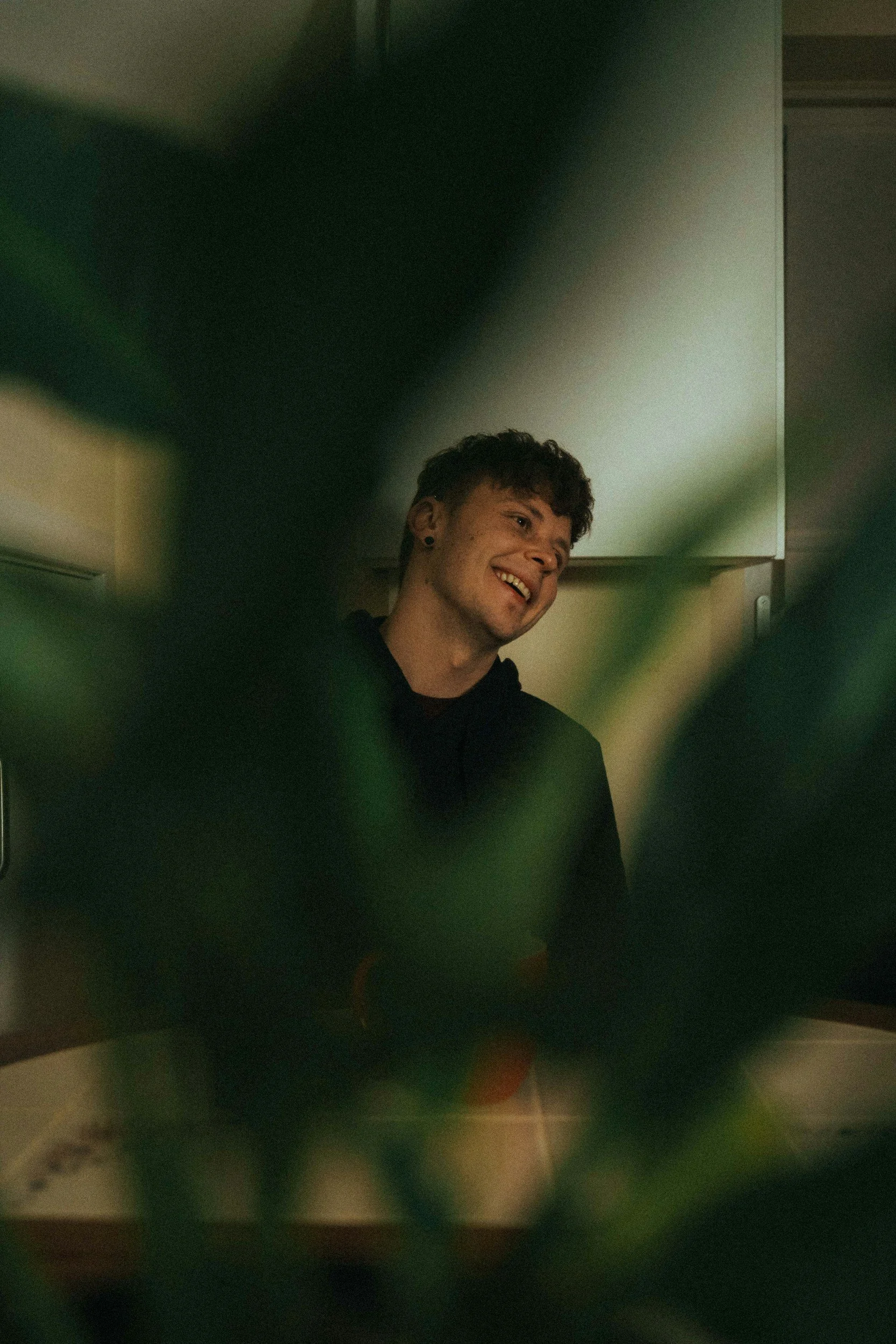 A young man with curly hair and earrings smiling, seen through some green leaves, in an indoor setting with dim lighting.