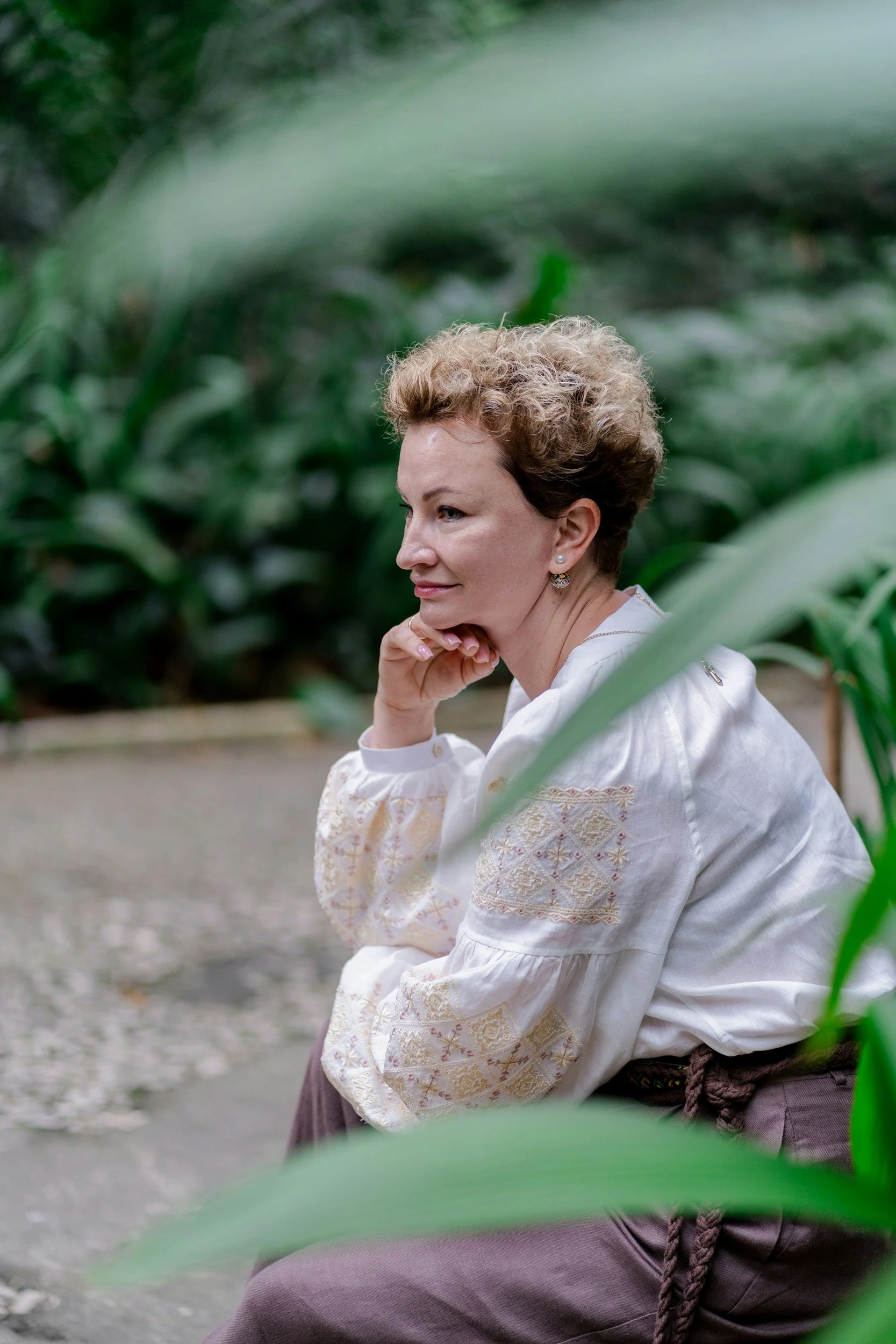 A woman with short, curly blonde hair sits outdoors among green plants, resting her chin on her hand, dressed in traditional clothing.