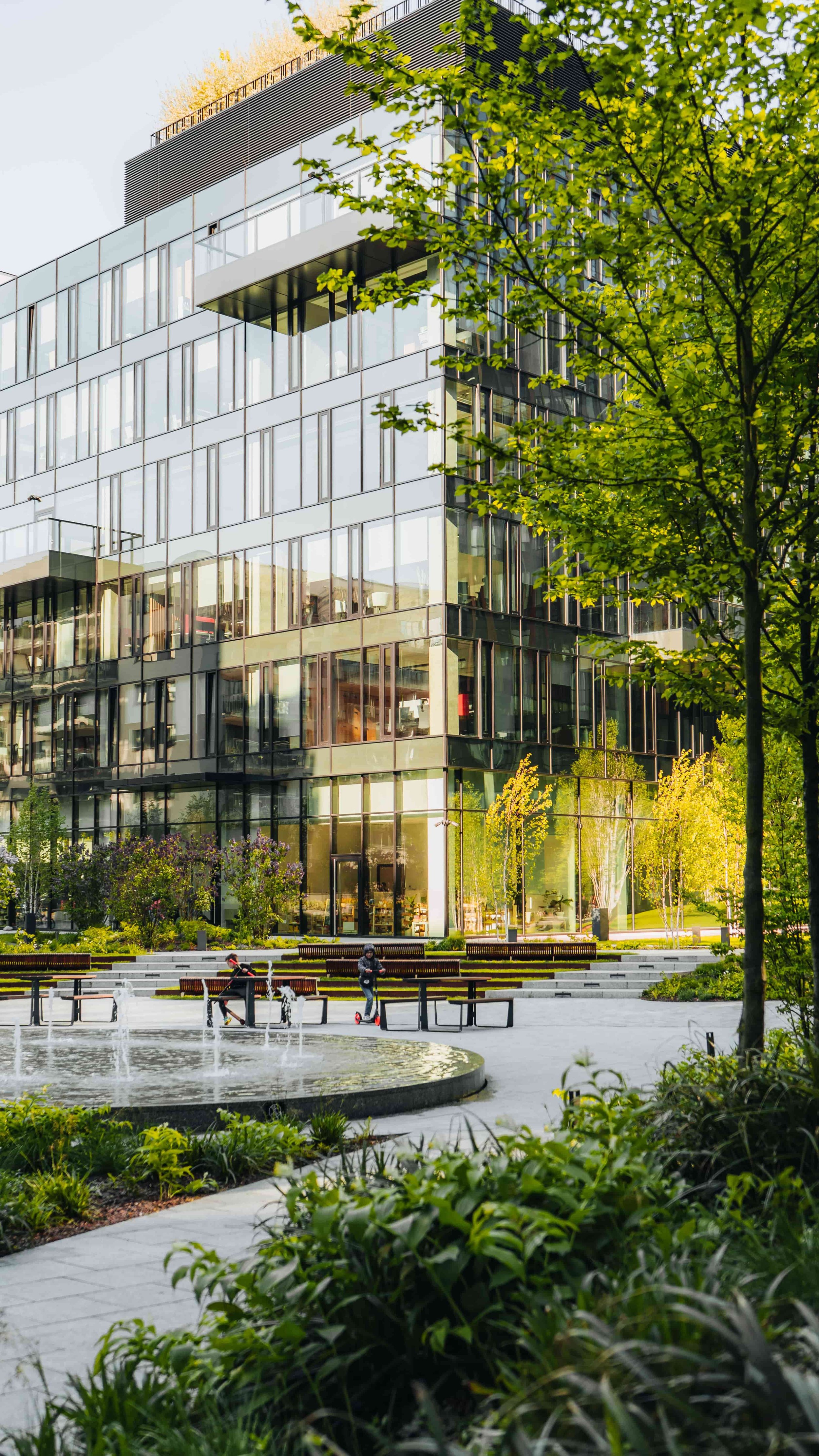 Modern glass building with greenery in foreground and people in the park.