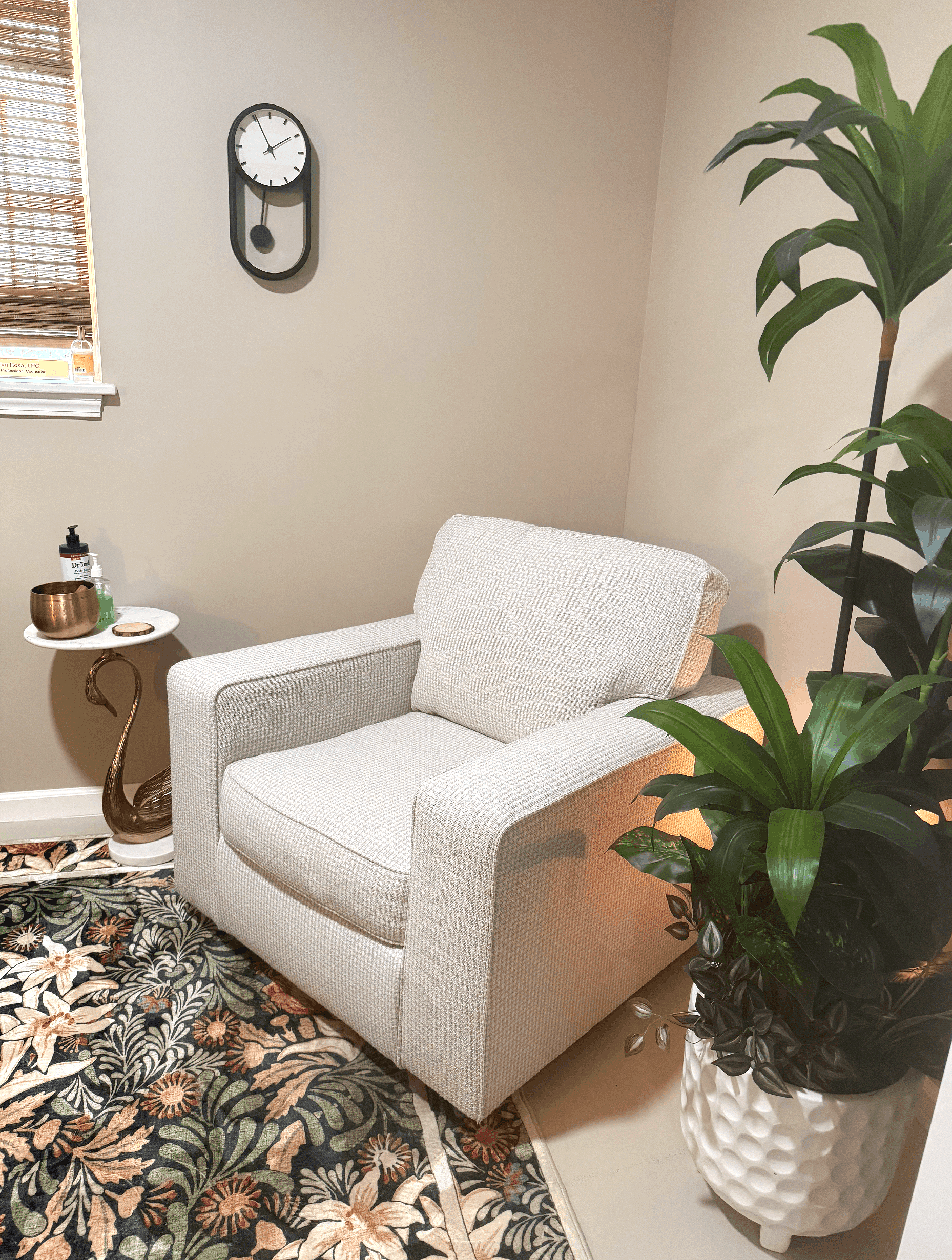 Interior of a cozy room with a white upholstered armchair, a small side table with decorative items, a large green houseplant in a textured white planter, and a wall clock. There's a window with wooden blinds and a patterned rug on the floor.