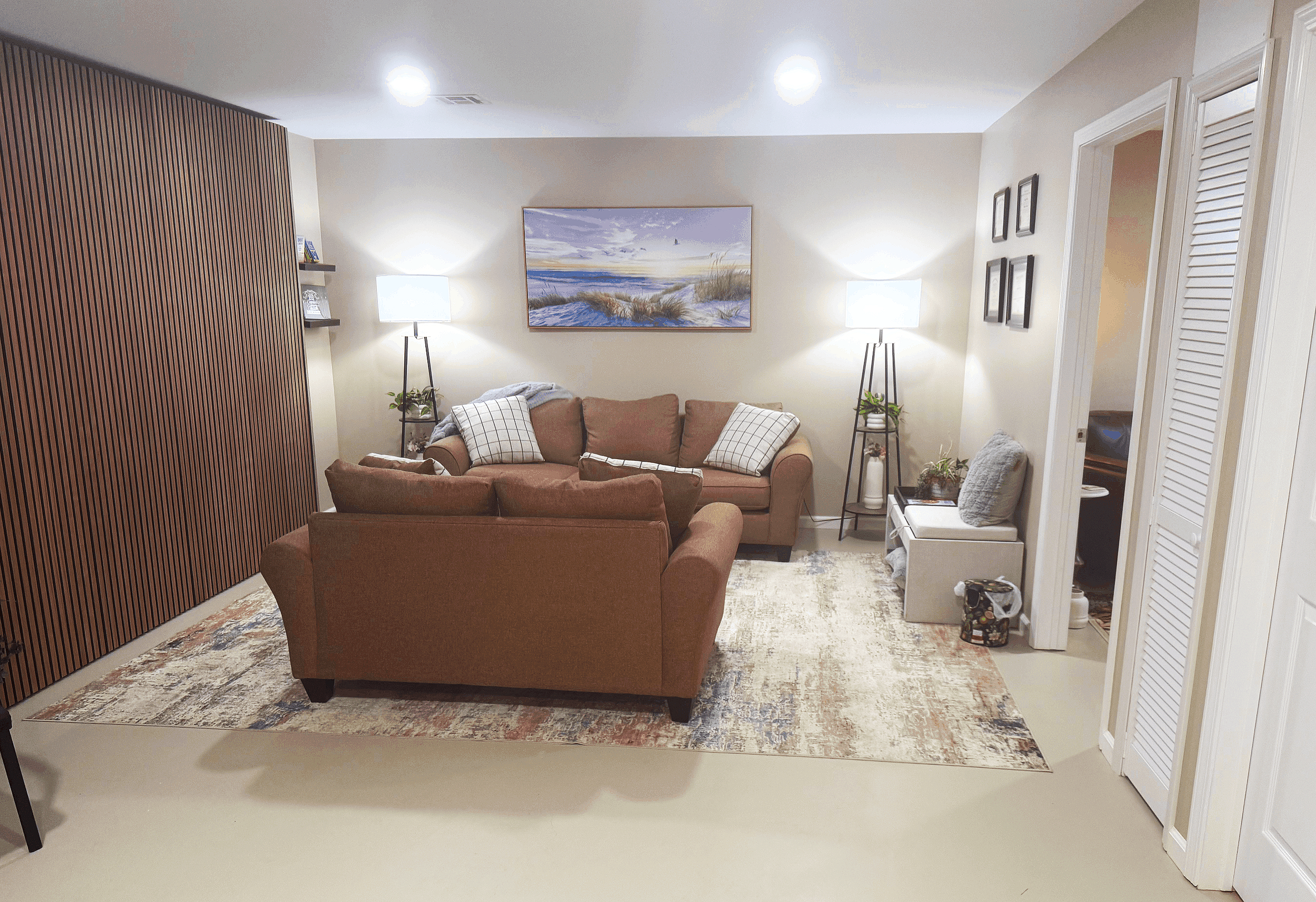 Living room with a brown sofa, two black side tables with lamps, a large landscape painting, and a wooden partition on the left.