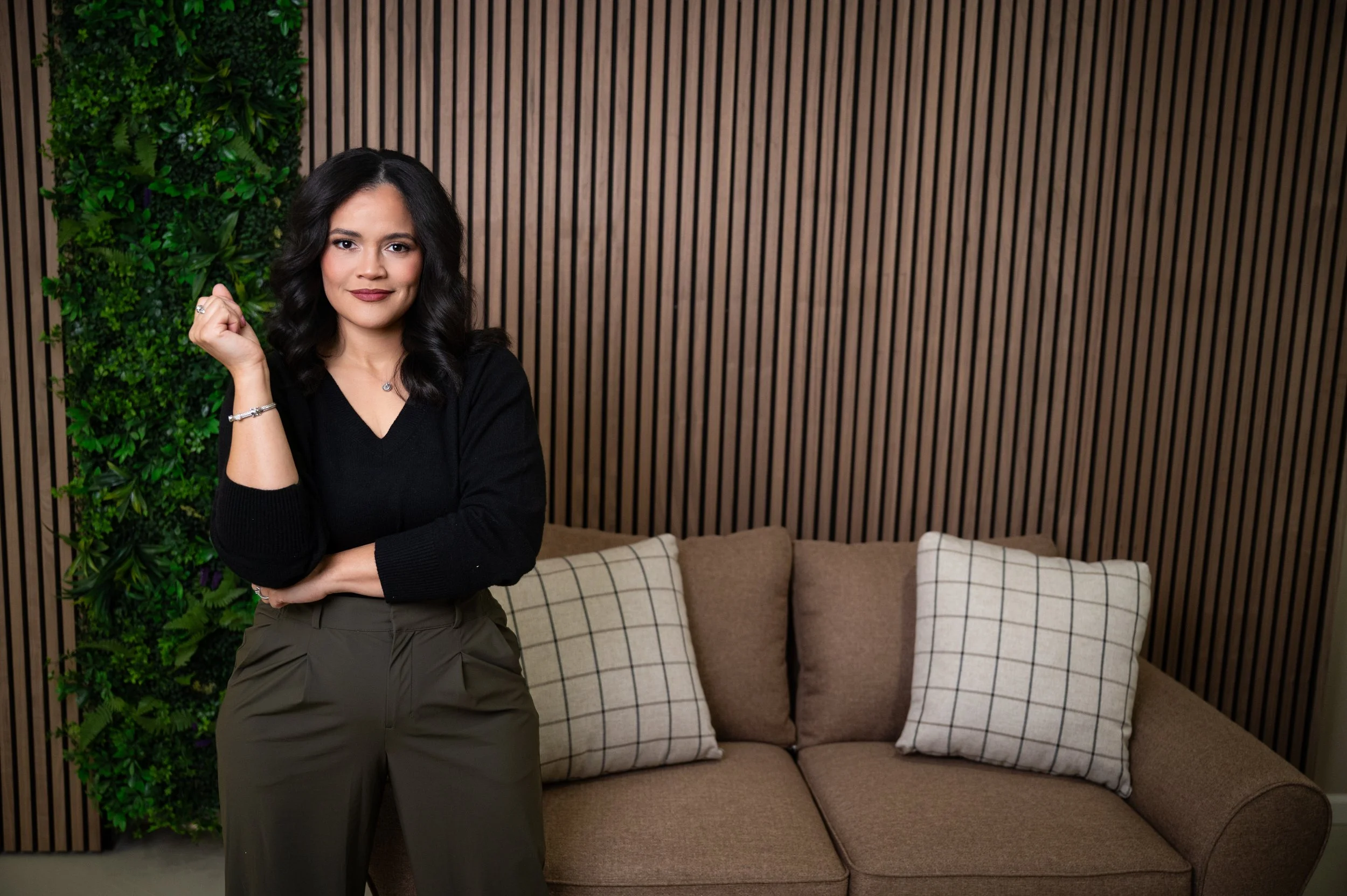 A woman with dark hair, wearing a black sweater and olive green pants, standing in front of a beige couch with plaid and plain cushions, next to a green vertical garden and wooden wall panels.