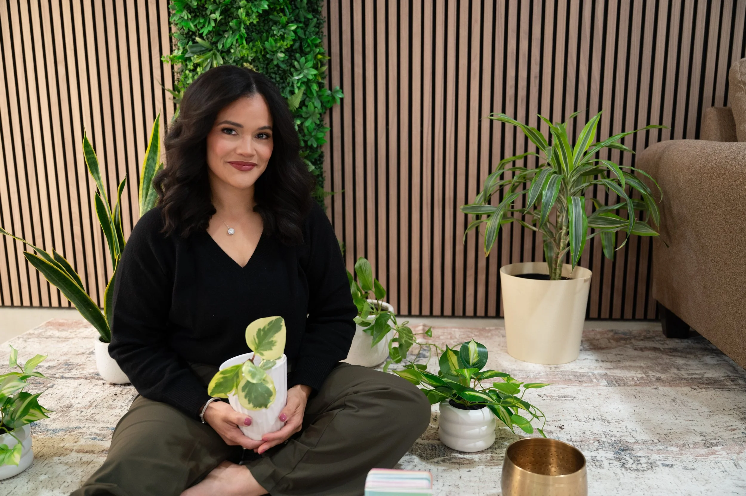 A woman sitting on a rug surrounded by potted green plants in an indoor setting, with a wooden slat wall in the background.