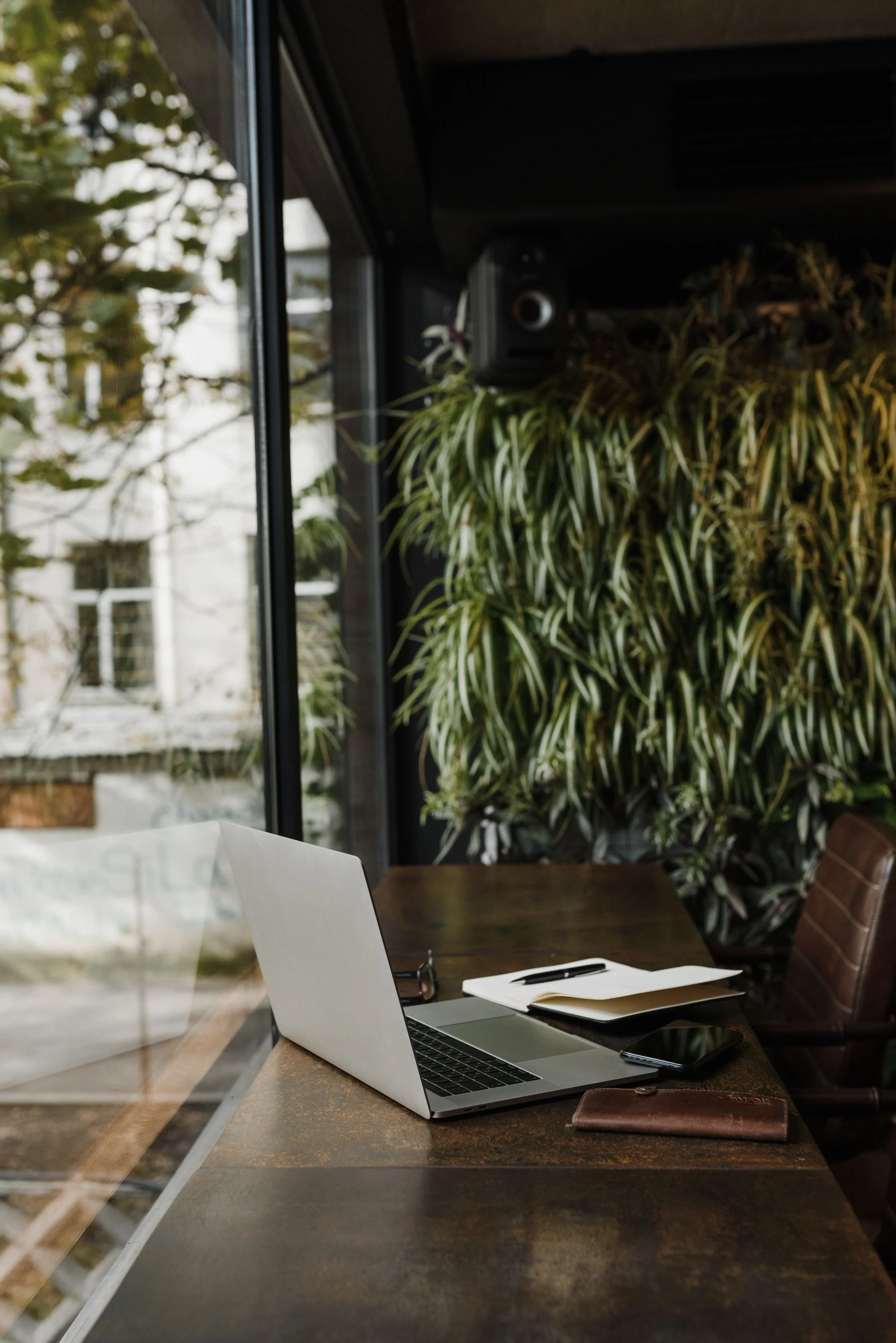 A laptop, a notebook with a pen, and a smartphone on a wooden table behind a glass window in a cozy cafe. In the background, there are green indoor plants and a large window showing an outdoor view with trees and a building.
