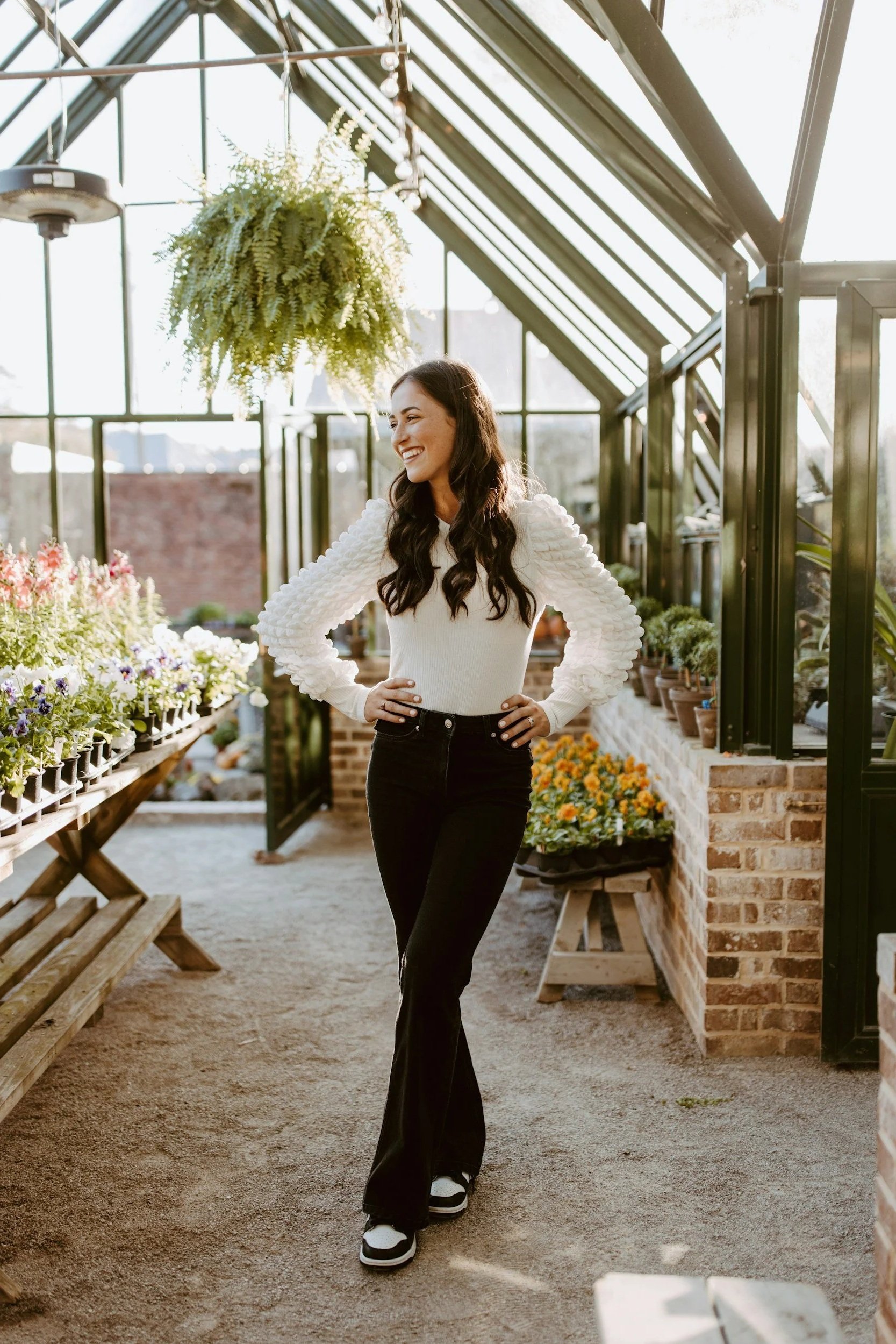 A woman with long dark hair smiling and standing inside a greenhouse filled with potted plants and flowers. She is wearing a white sweater, black pants, and sneakers.