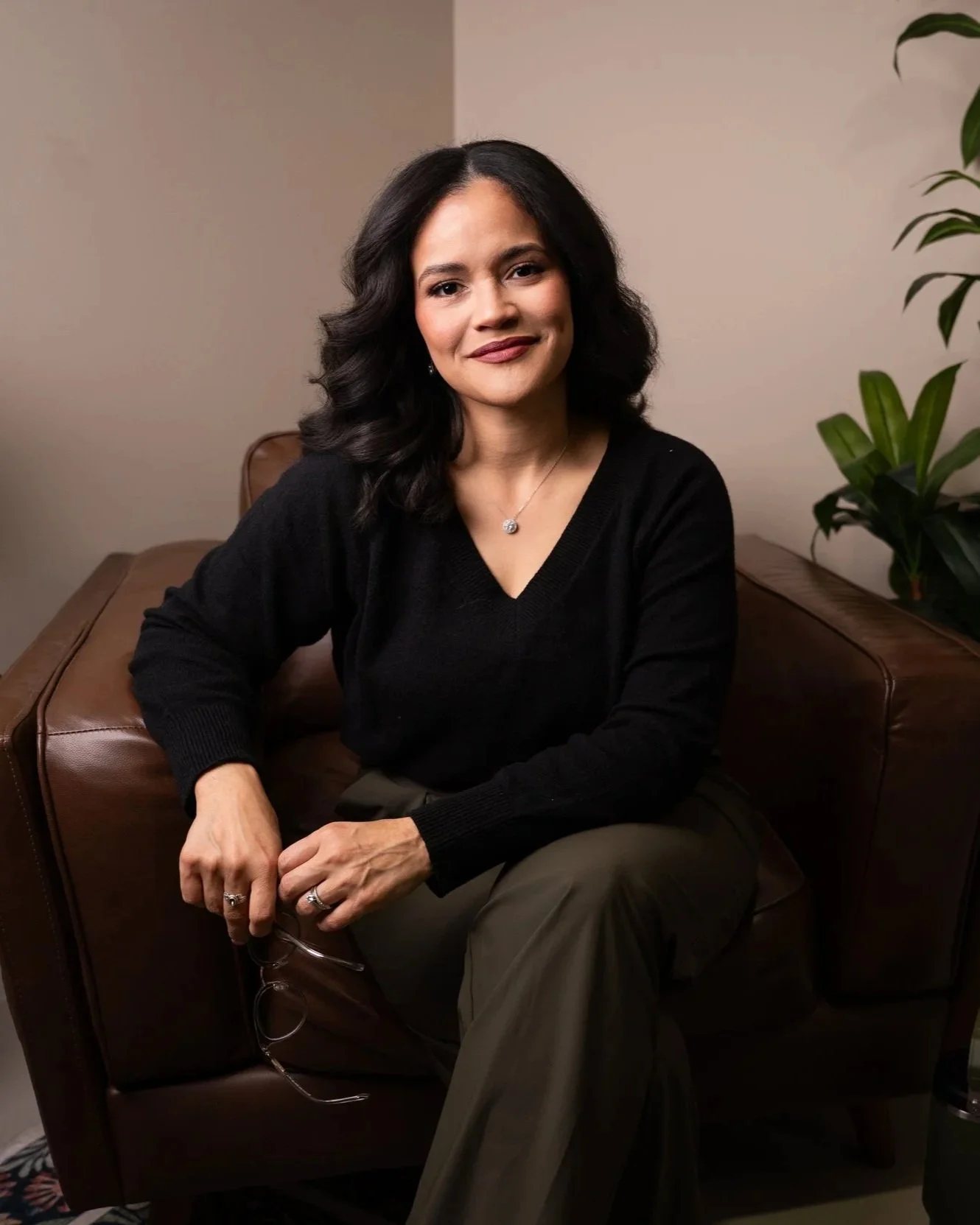 A woman with dark wavy hair wearing a black sweater and jewelry sitting on a brown leather couch in a room with neutral-colored walls and a plant.