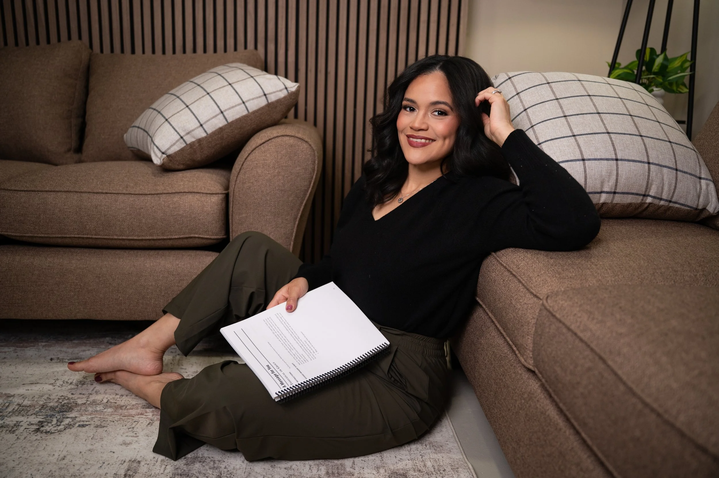 A smiling woman with dark hair, wearing a black sweater and green pants, sitting on the floor next to a brown couch with her back against an armrest, holding a spiral-bound document in her lap. The couch has a plaid pillow, and a small green plant is on a side table in the background.