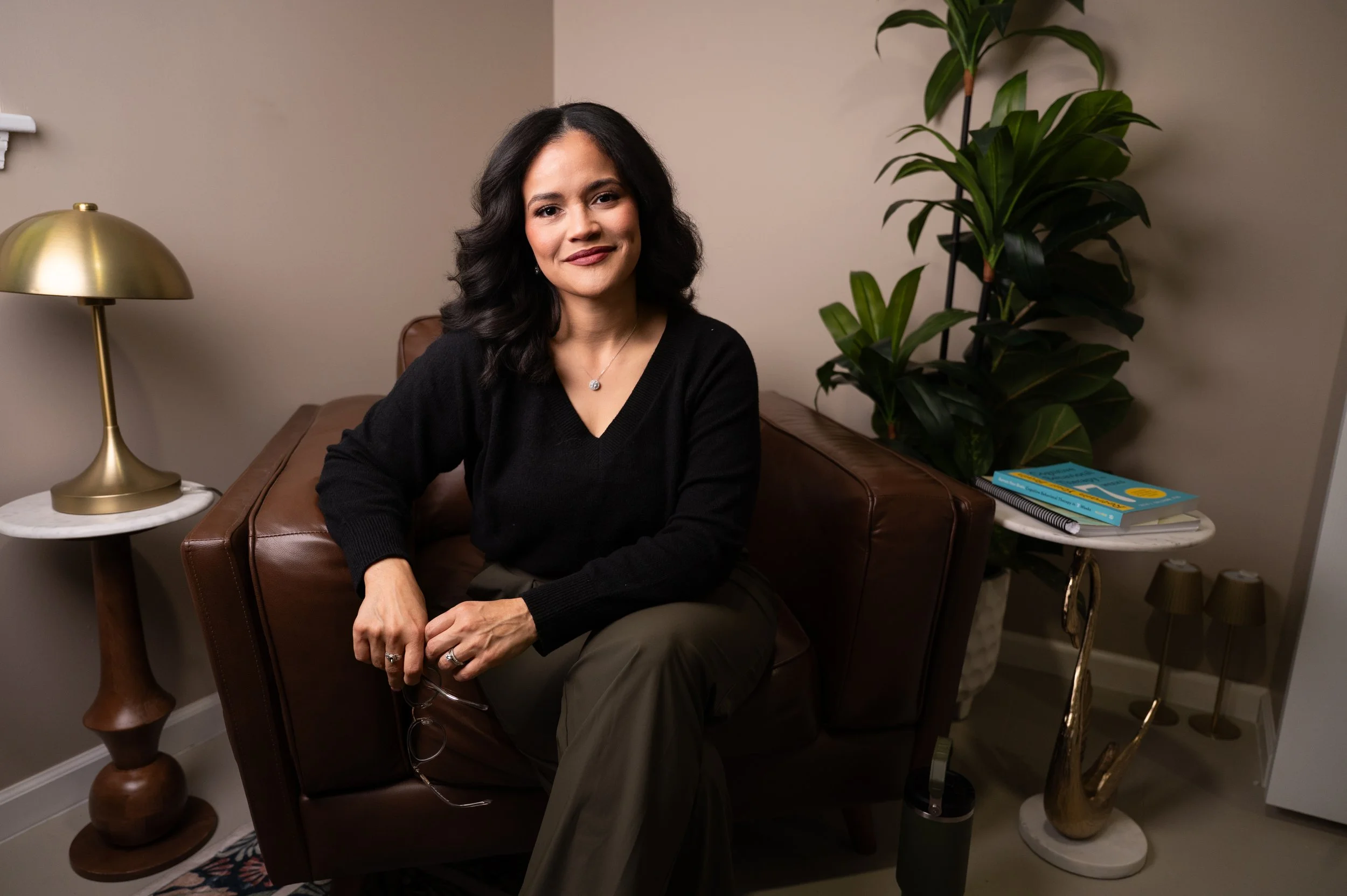 A woman with black wavy hair, wearing a black sweater and khaki pants, sitting on a brown leather armchair in a cozy room with beige walls, next to a white side table with a gold lamp, and a plant with green leaves. She is smiling at the camera, holding eyeglasses, and wearing a necklace.