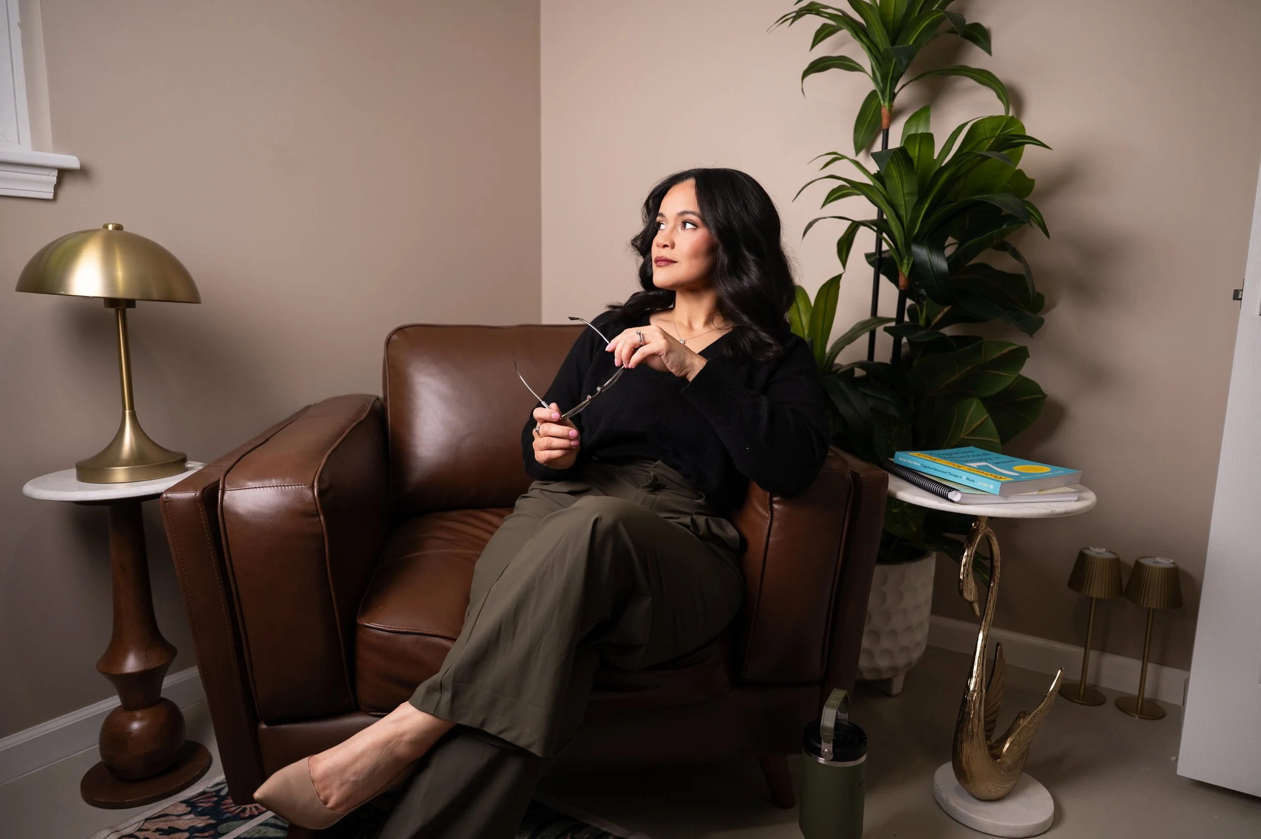 Woman with dark hair sitting on a leather sofa, holding glasses, in a cozy, modern living room with a gold lamp, large green plant, and decorative items.