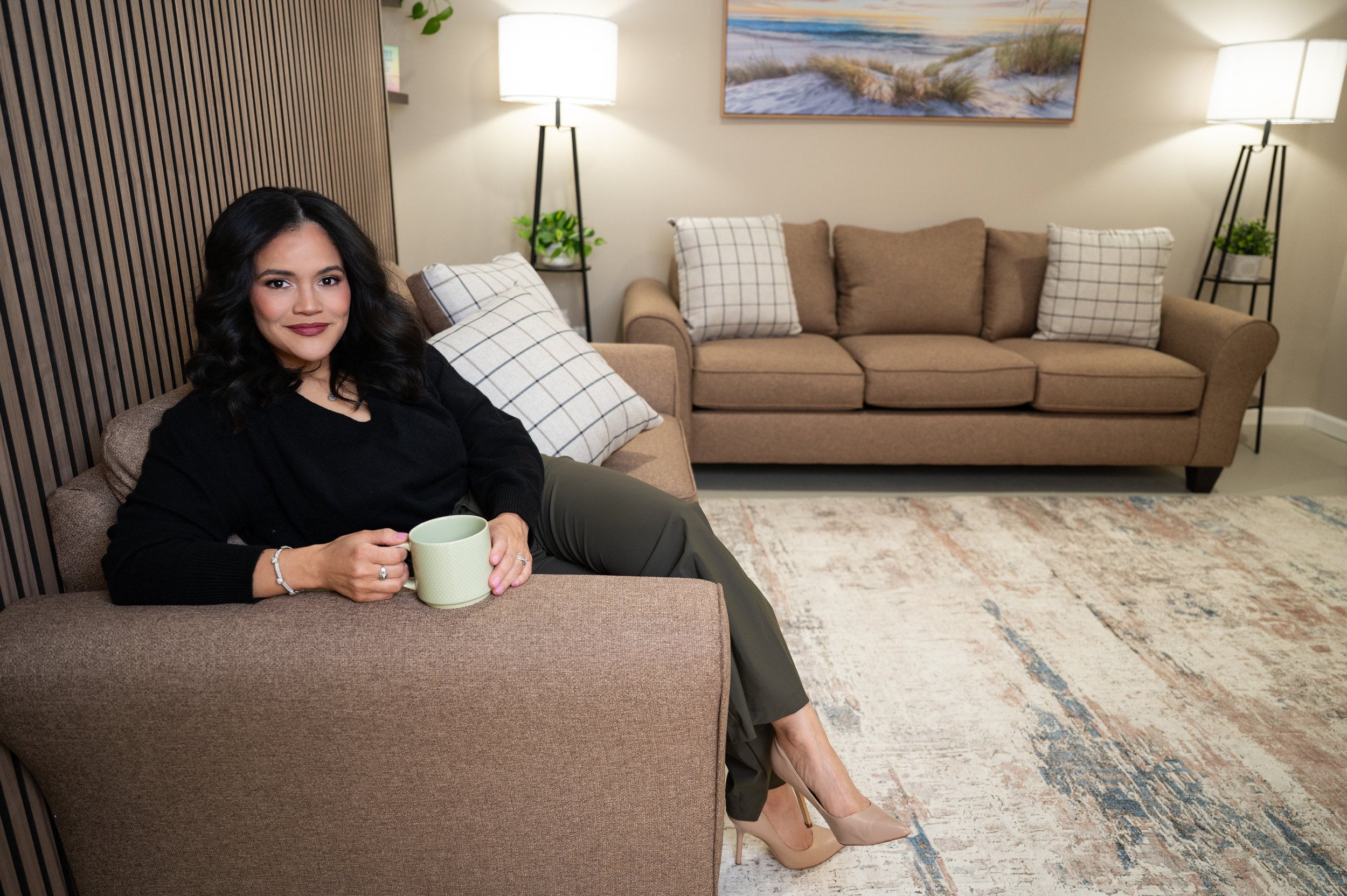 A woman with dark wavy hair sitting on a beige sofa in a living room, holding a white mug, with a neutral expression, wearing a black sweater and olive-green pants, beige high heels, and a bracelet, surrounded by pillows and modern lamps.