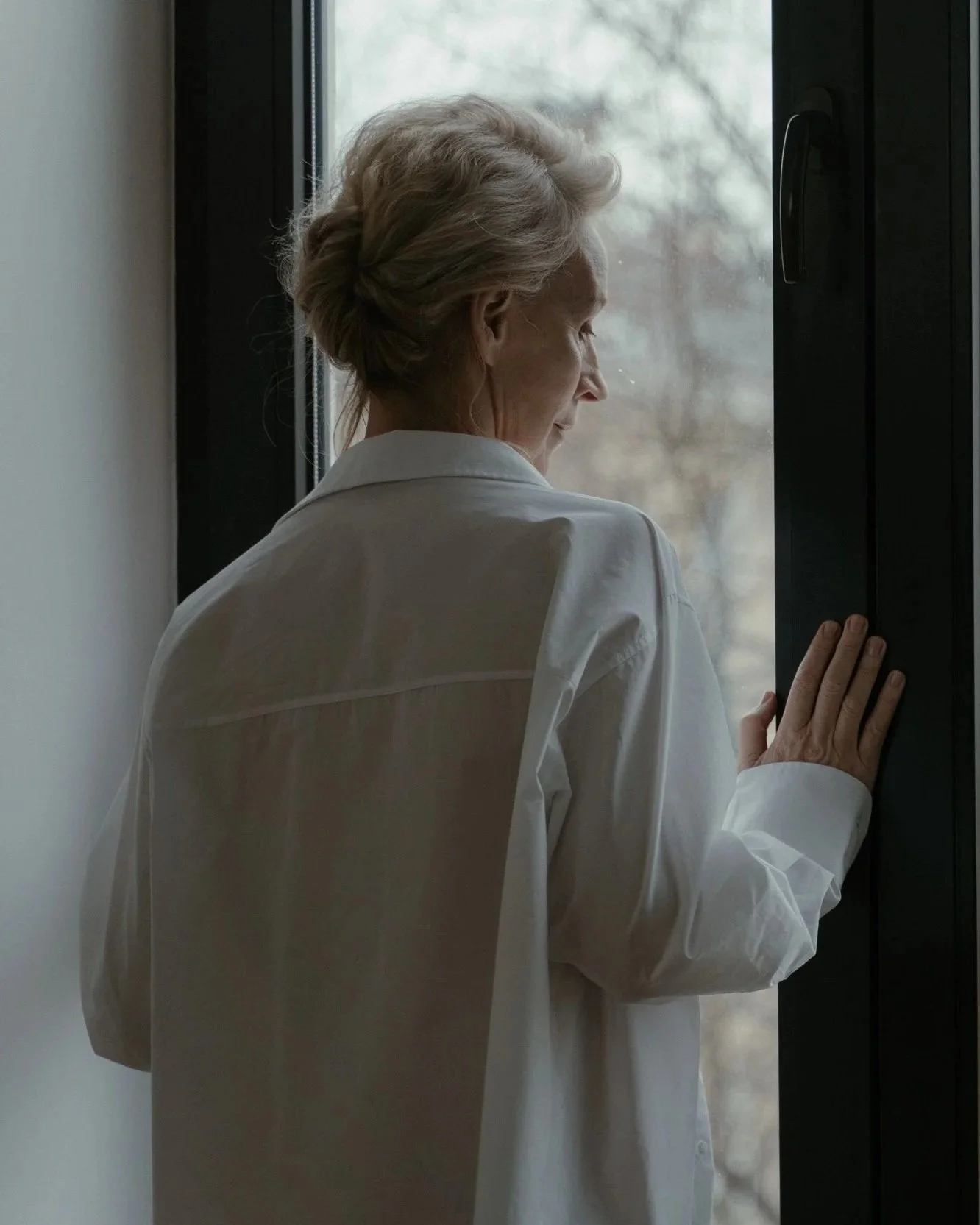 An elderly woman with gray hair wearing a white shirt, looking out the window and gently touching the window frame with her hand.