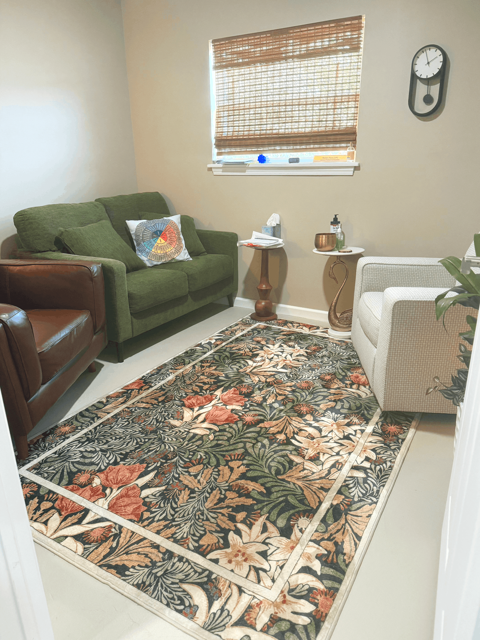 A living room with a patterned area rug on the floor, a green sofa with pillows, a side table with tissues and decorative items, a window with blinds, and a white armchair. Wall clock is on the wall.