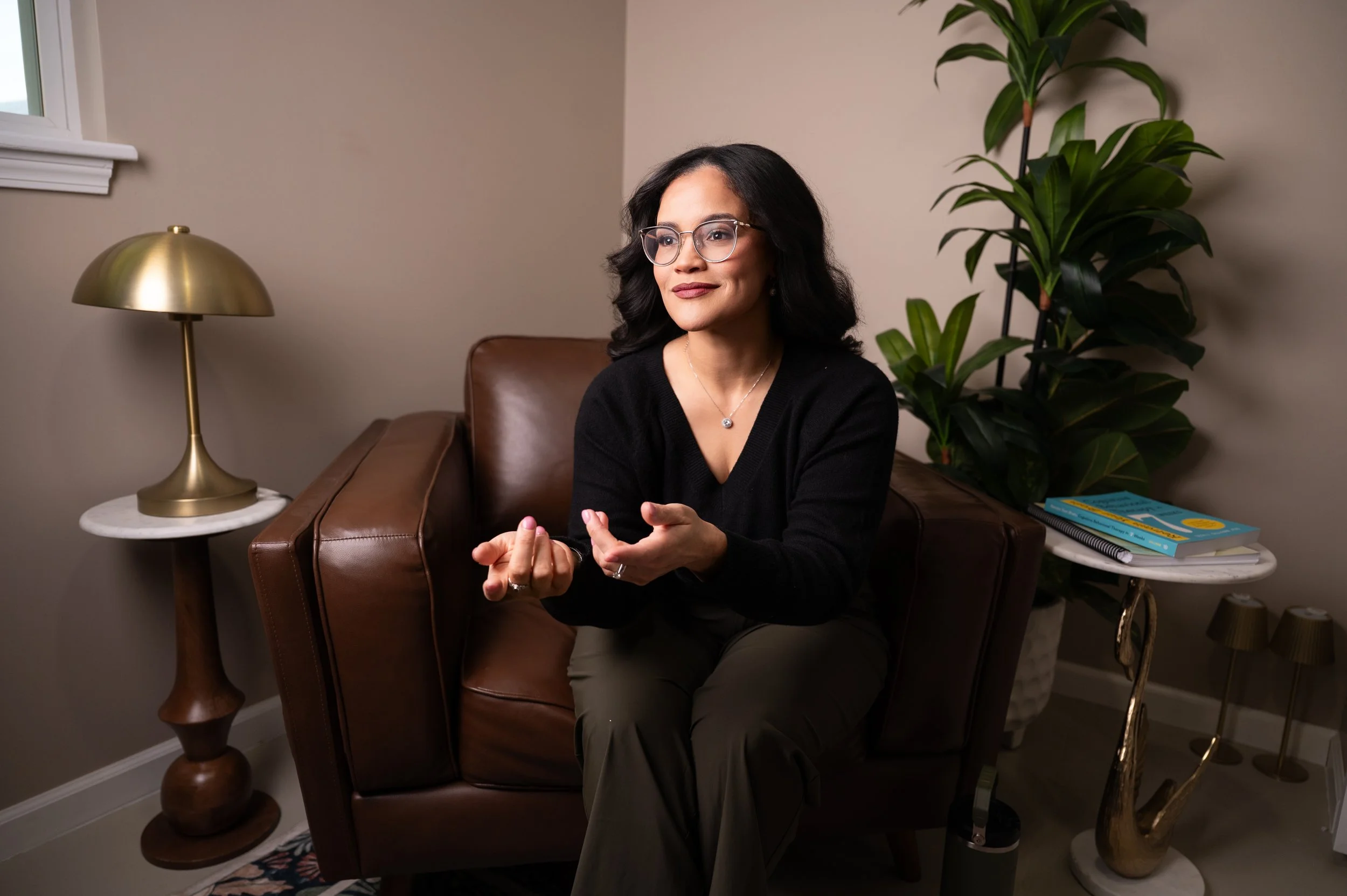 A woman with dark hair, glasses, and a black sweater sitting on a brown leather sofa in a cozy room, gesturing with her hands, with a gold and white table lamp, green potted plant, and a side table with books in the background.