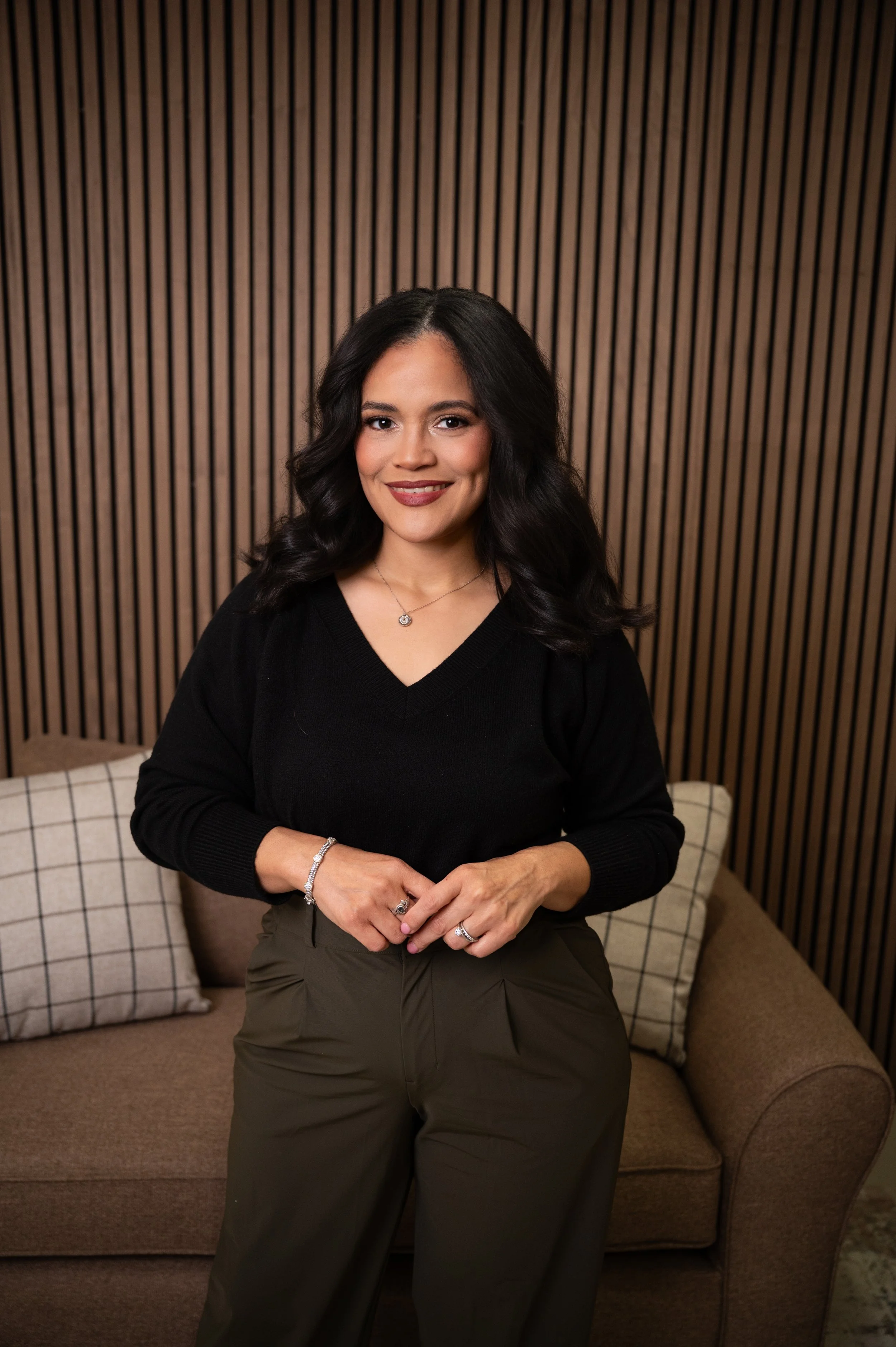 A woman with dark wavy hair, wearing a black sweater, olive green pants, a necklace, rings, and a bracelet, sitting on a brown sofa in front of a wooden slat wall.