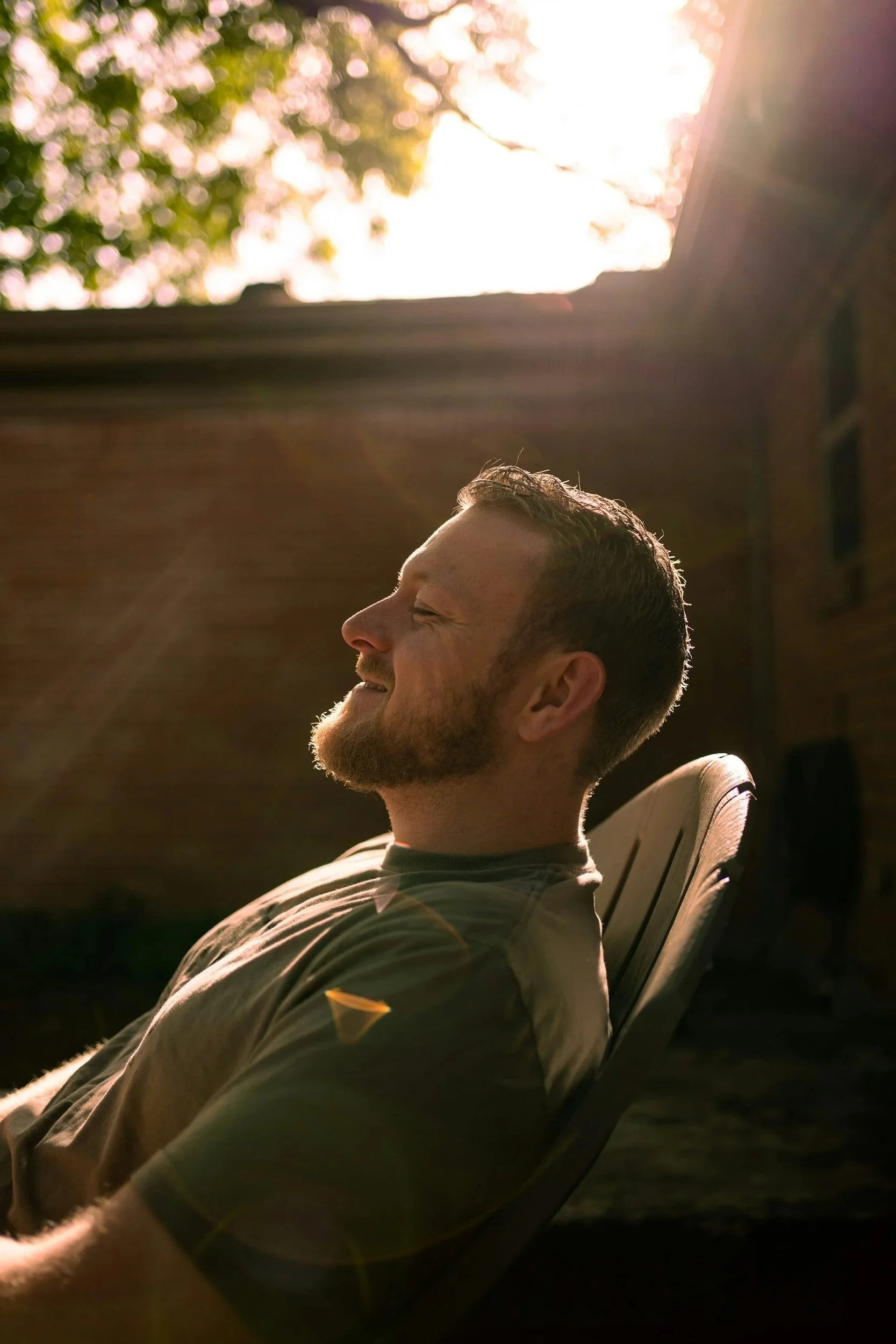 A man is sitting outdoors on a lounge chair, smiling and relaxing in the sunlight with his eyes closed, with trees visible in the background and the sun shining brightly.