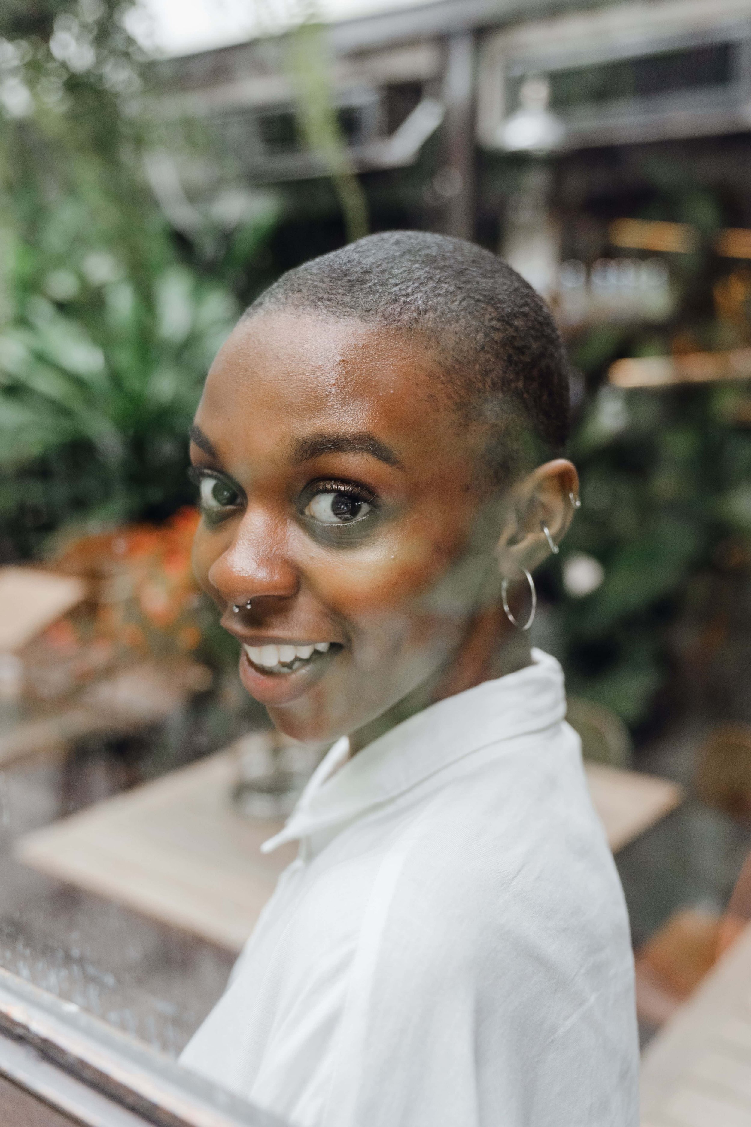 Close-up of a smiling young woman with short natural hair looking over her shoulder, wearing earrings and a white shirt, with a blurred background of lush green plants.
