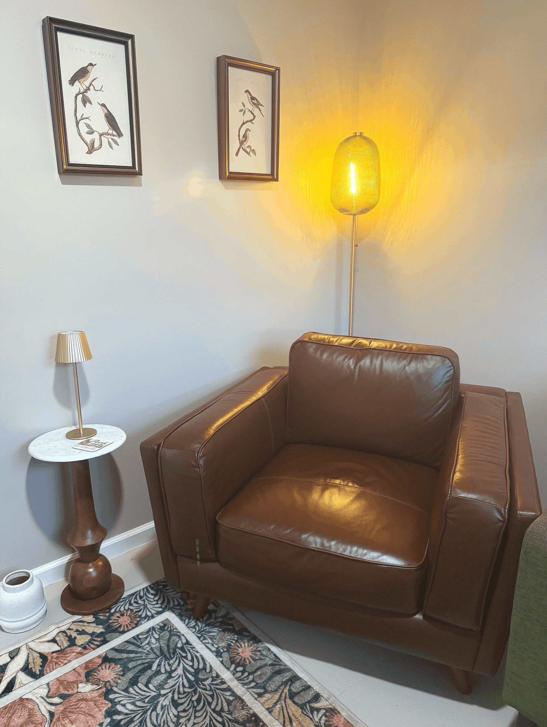A living room corner with a brown leather armchair, a side table with a white marble top, a small beige lamp, a decorative vase, a colorful floral rug, and wall art featuring framed bird illustrations and a yellow hanging lamp illuminating the space.
