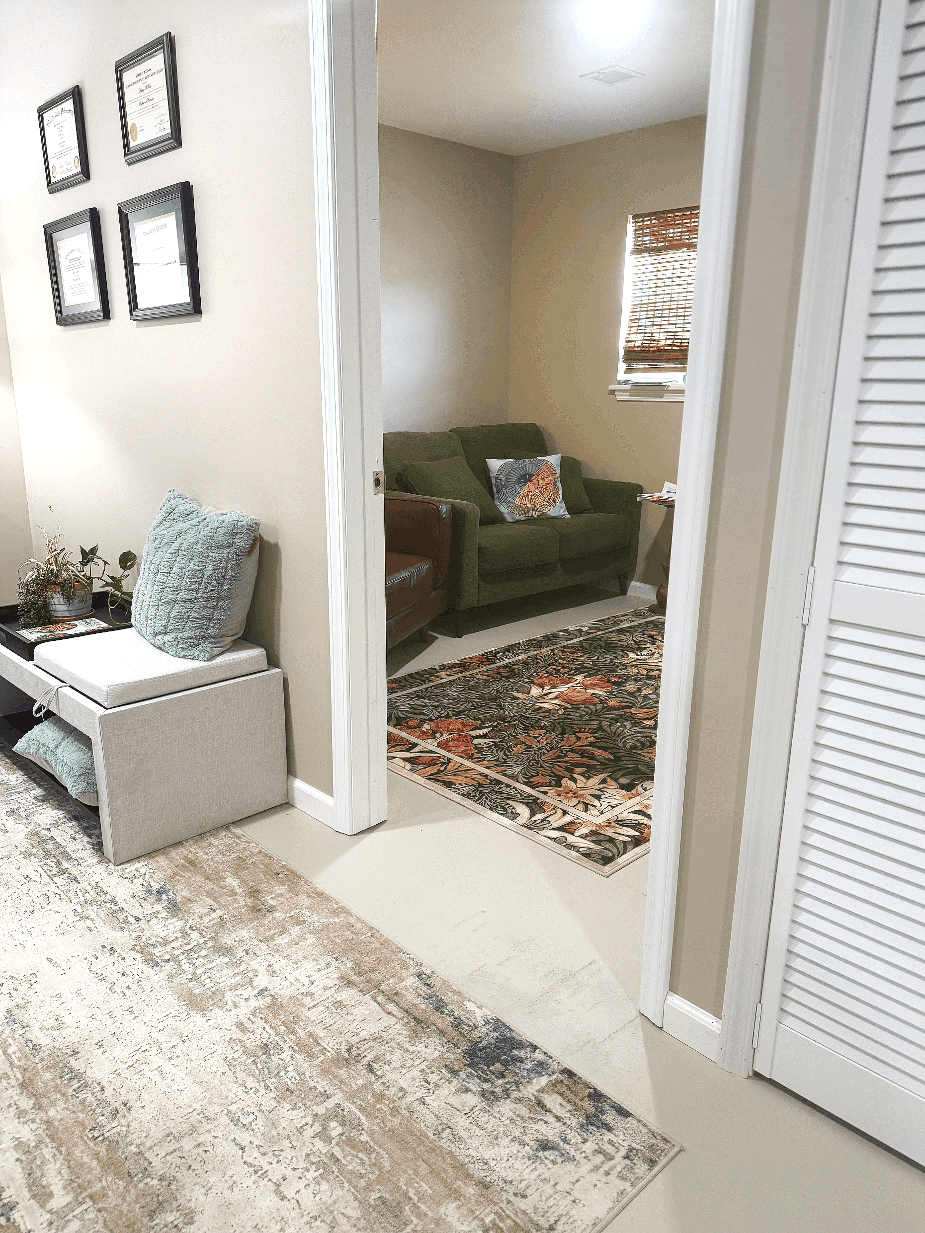 View of a room with a patterned rug, white wall with framed certificates, beige wall with a window, green couch with pillows, partially open white louvered closet door, and a small white table with a pillow and plant on it.