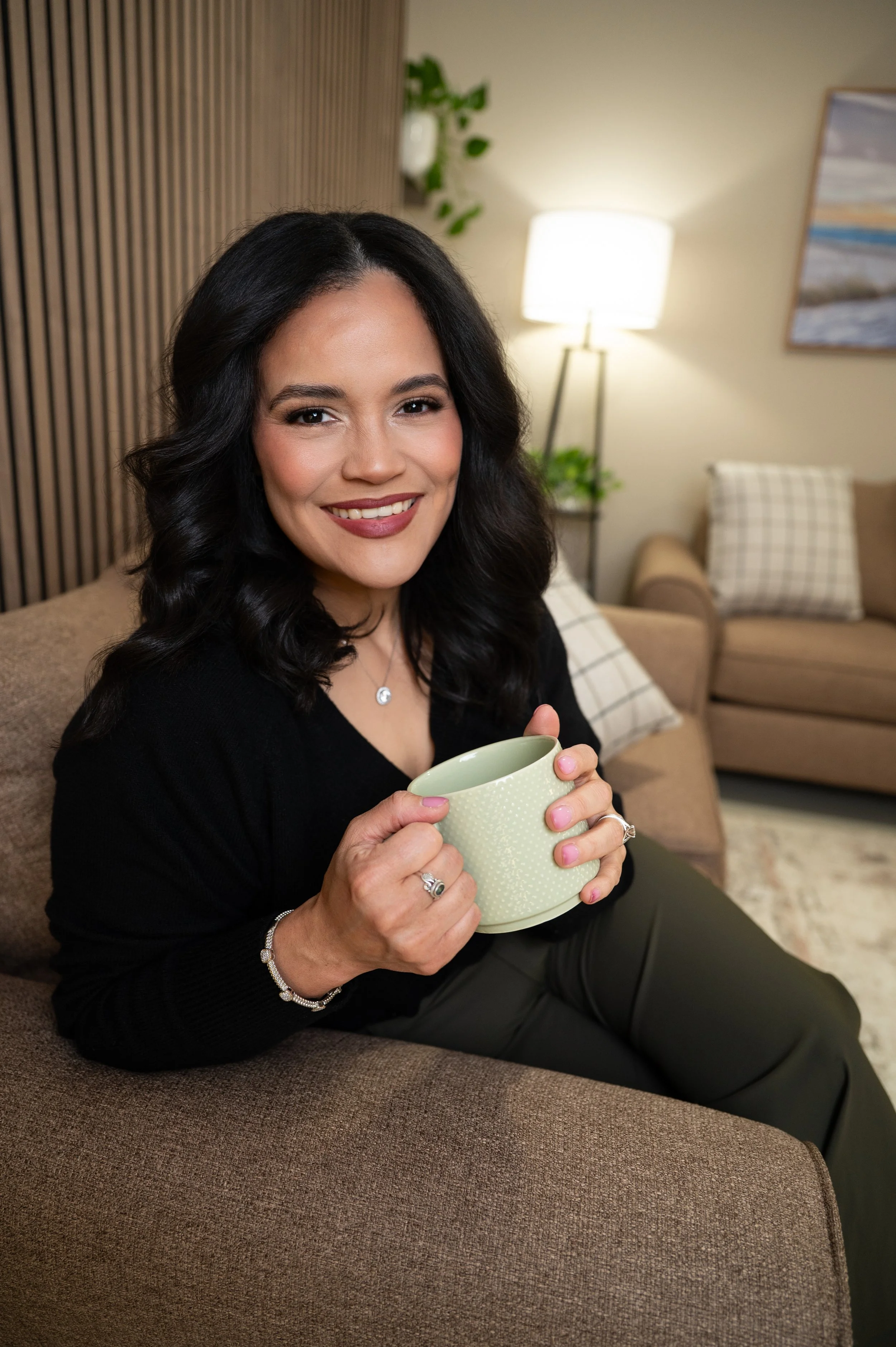 A woman with dark, wavy hair and a black top sitting on a brown sofa in a cozy living room, holding a light green mug and smiling at the camera.