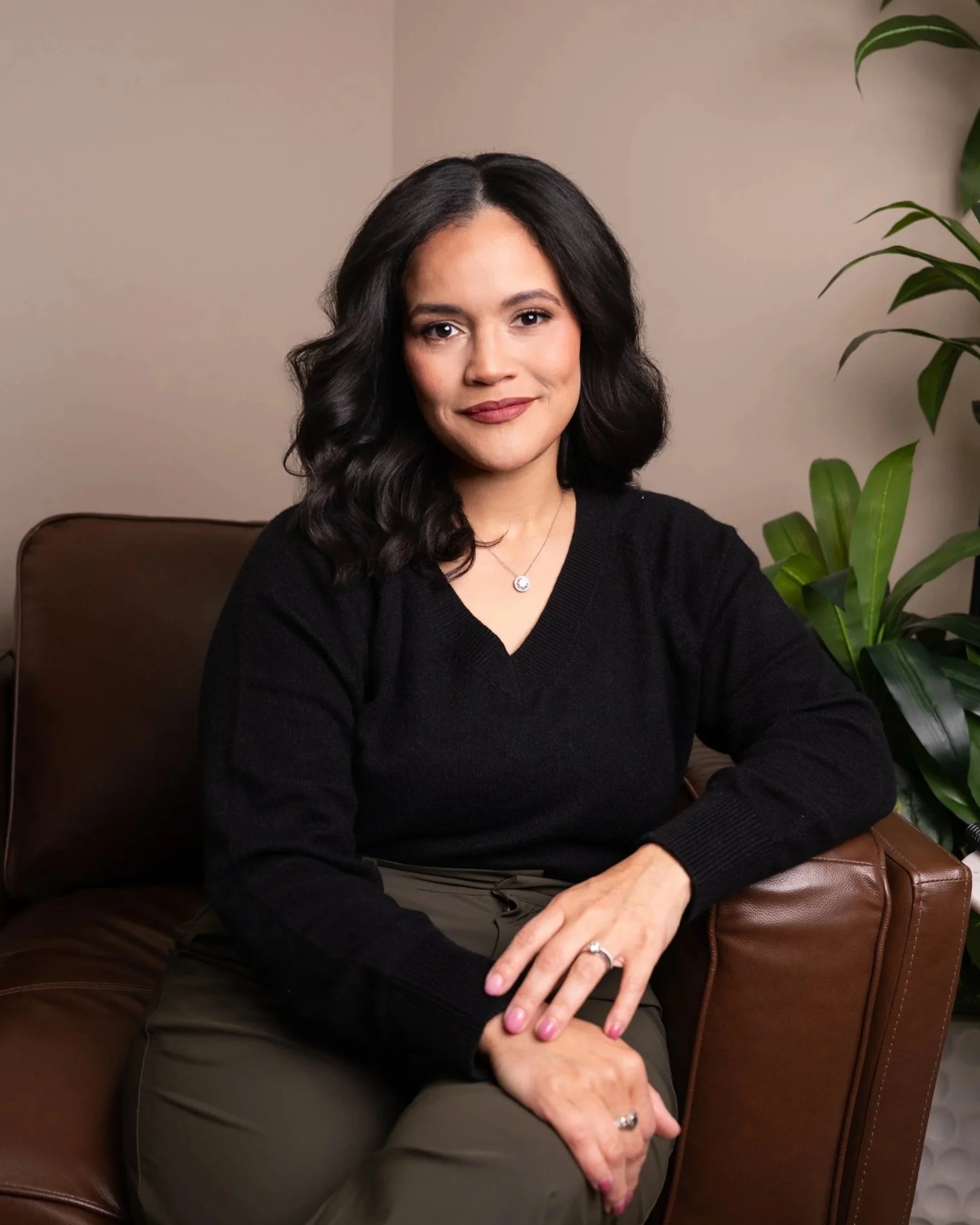 A woman with black, wavy hair, wearing a black sweater and gold necklace, sitting on a brown leather couch next to a green houseplant, with beige walls in the background.