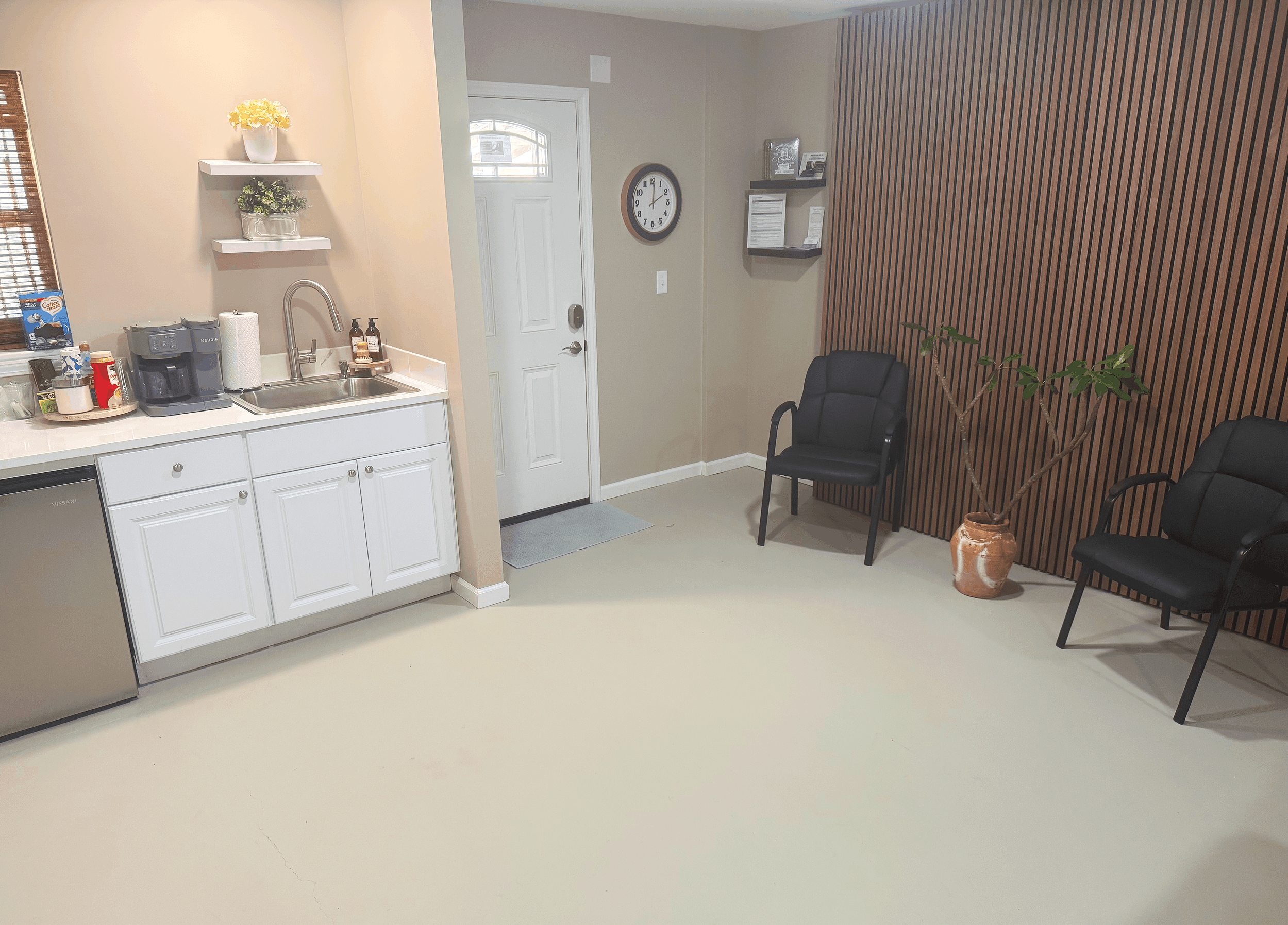 Interior of a room with a kitchenette on the left and a seating area on the right. The kitchenette features white cabinets, a small sink, a coffee maker, and shelves with decorative items. The seating area has black chairs, a potted plant, and decorative wall shelves. The room has a beige wall with vertical wood paneling and a clock near the door.