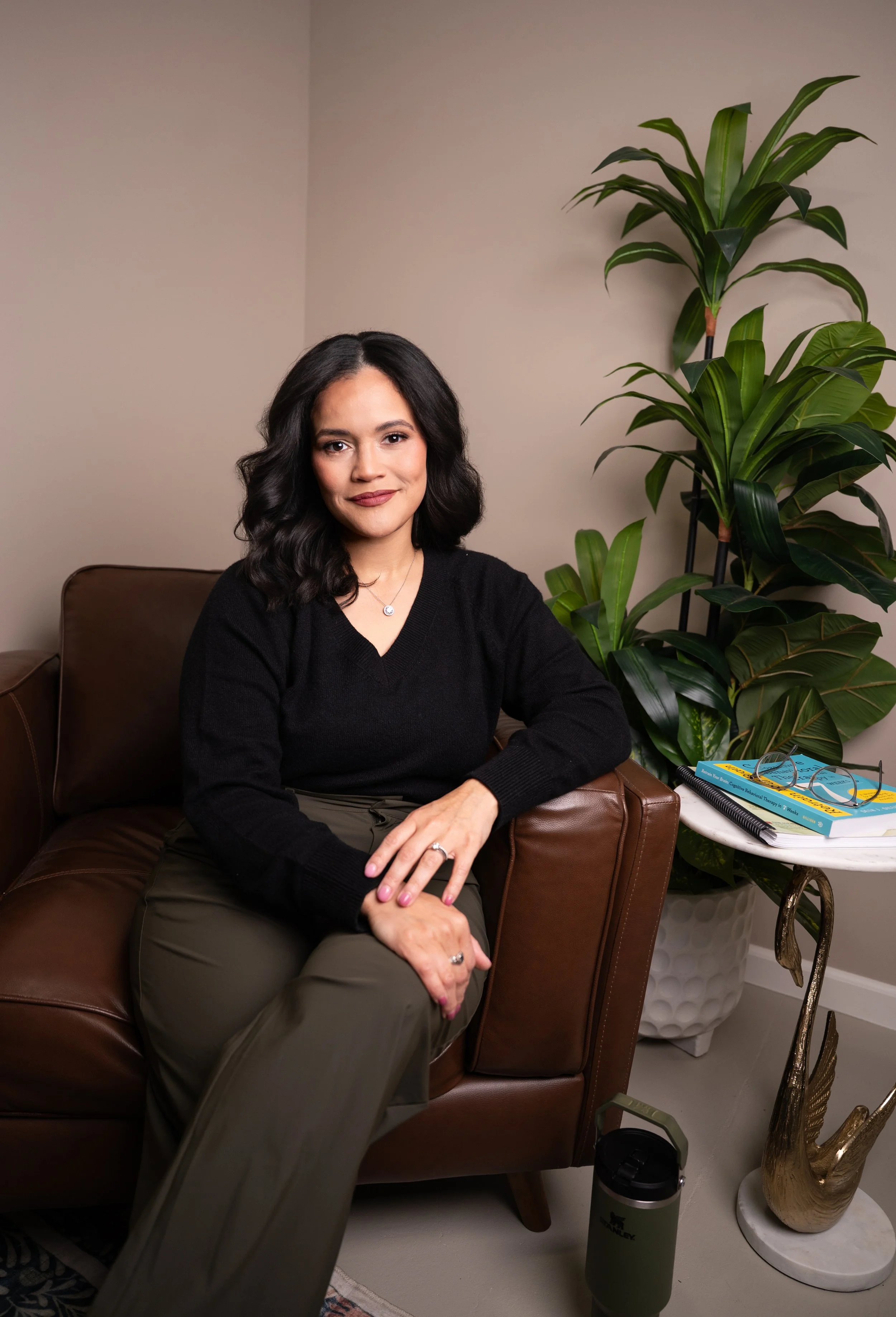 A woman sitting on a brown leather sofa with a confident smile, wearing a black sweater and khaki pants, in a room with a large green plant, a white side table with books, and decorative items.