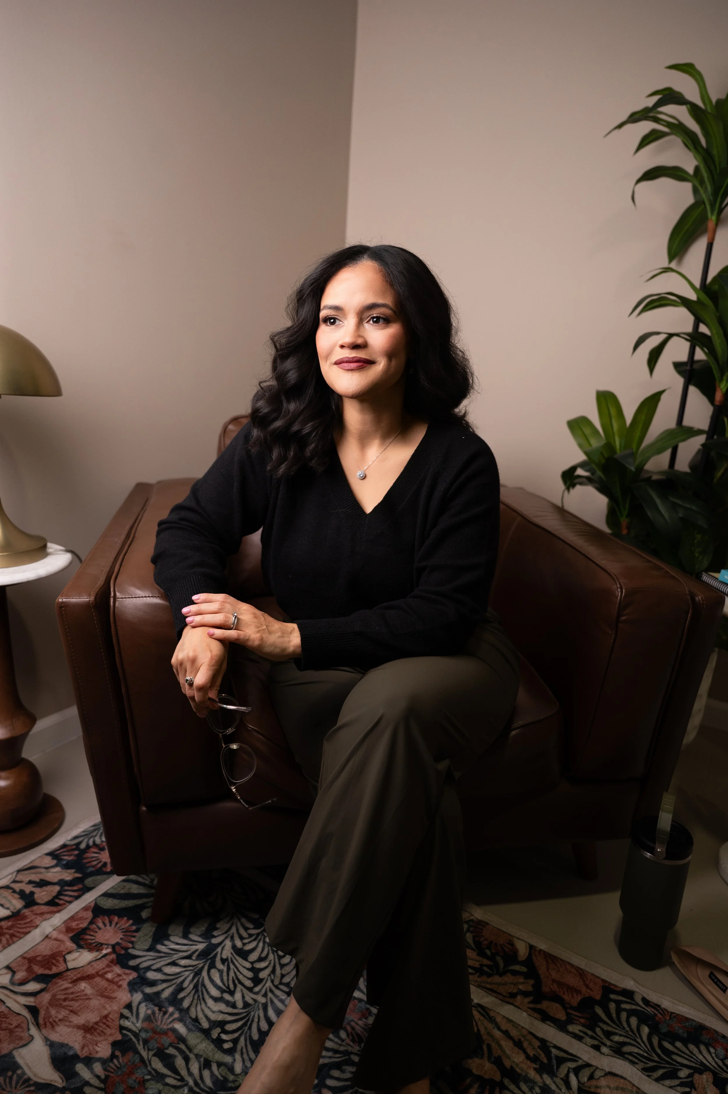 A woman with black wavy hair, wearing a black sweater and olive green pants, seated on a brown leather armchair in a cozy room with a beige wall, a green potted plant, a gold lamp on a side table, and a decorative rug.