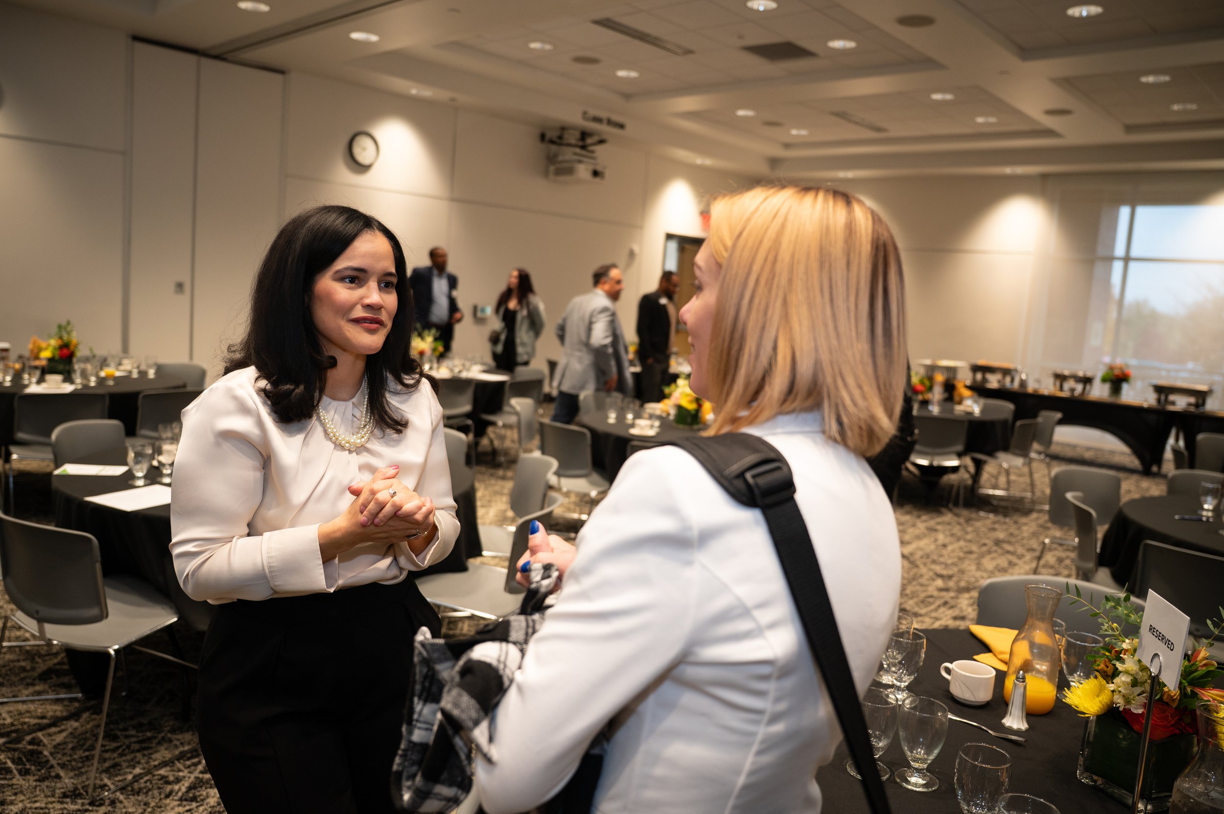 Two women engaged in a conversation at a formal event or conference, with tables set with glassware and floral arrangements in a large, well-lit room.