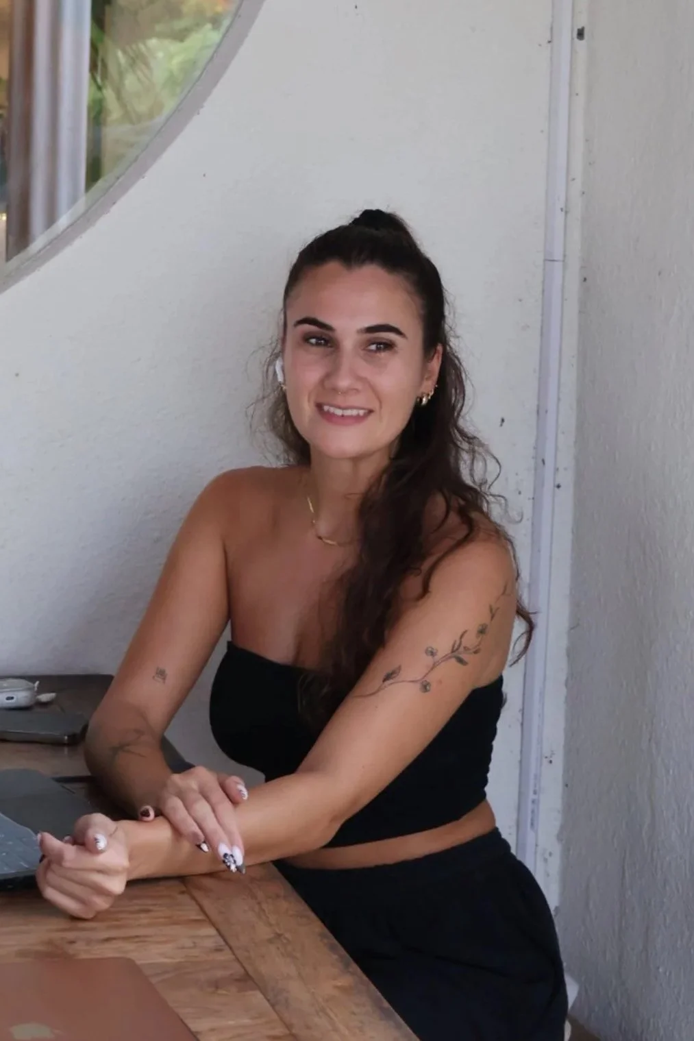 white woman with tanned skin, brown hair tied half up and half down wearing a black strapless top. tattoo of flowers on her left arm, looking ahead of the camera with a gentle smile and her hands on the table