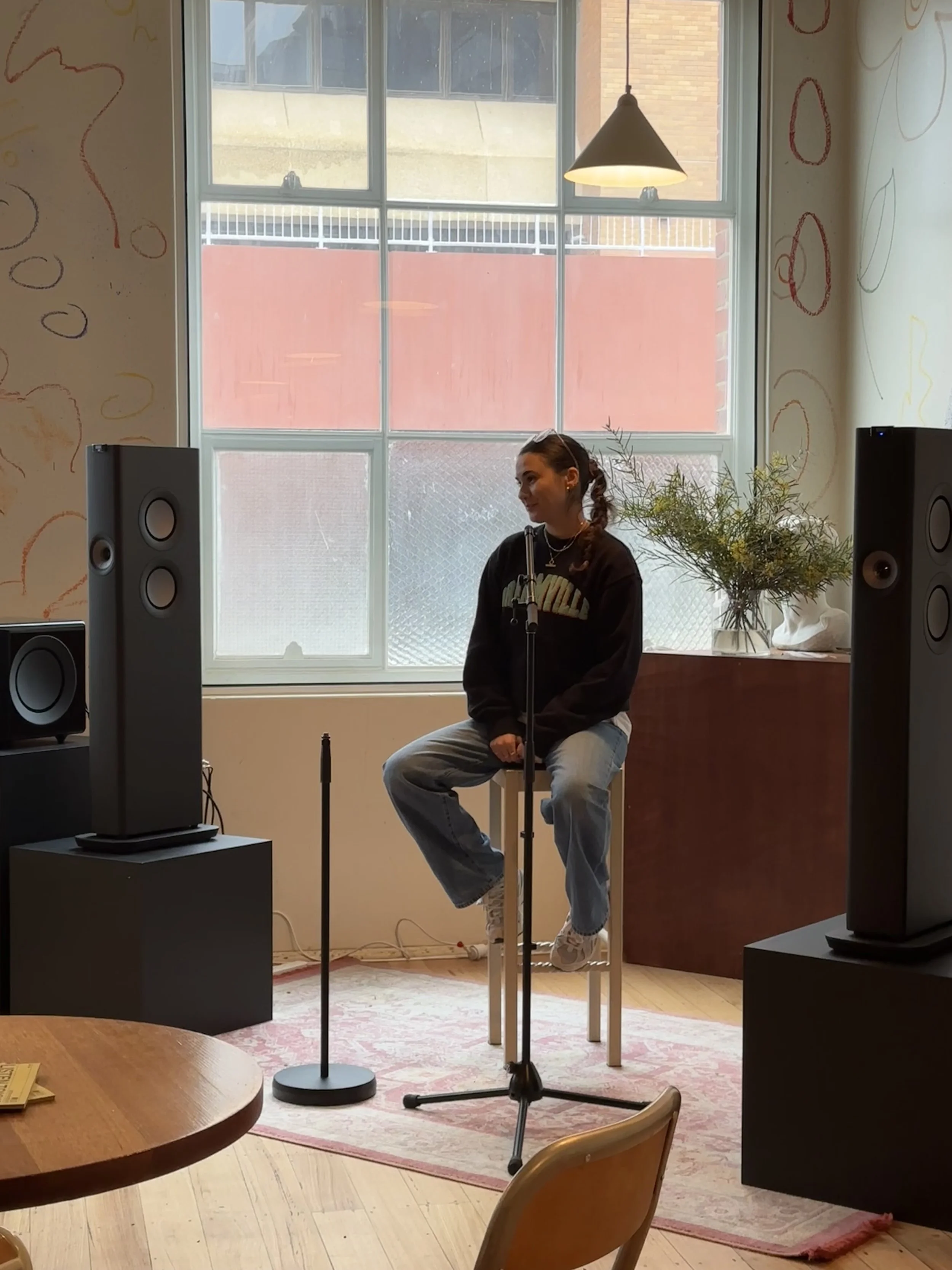 lizzy sat on a high stool wearing a black jumper and blue jeans, in between two KEF LS60 Wireless black speakers with a microphone stand in front of her