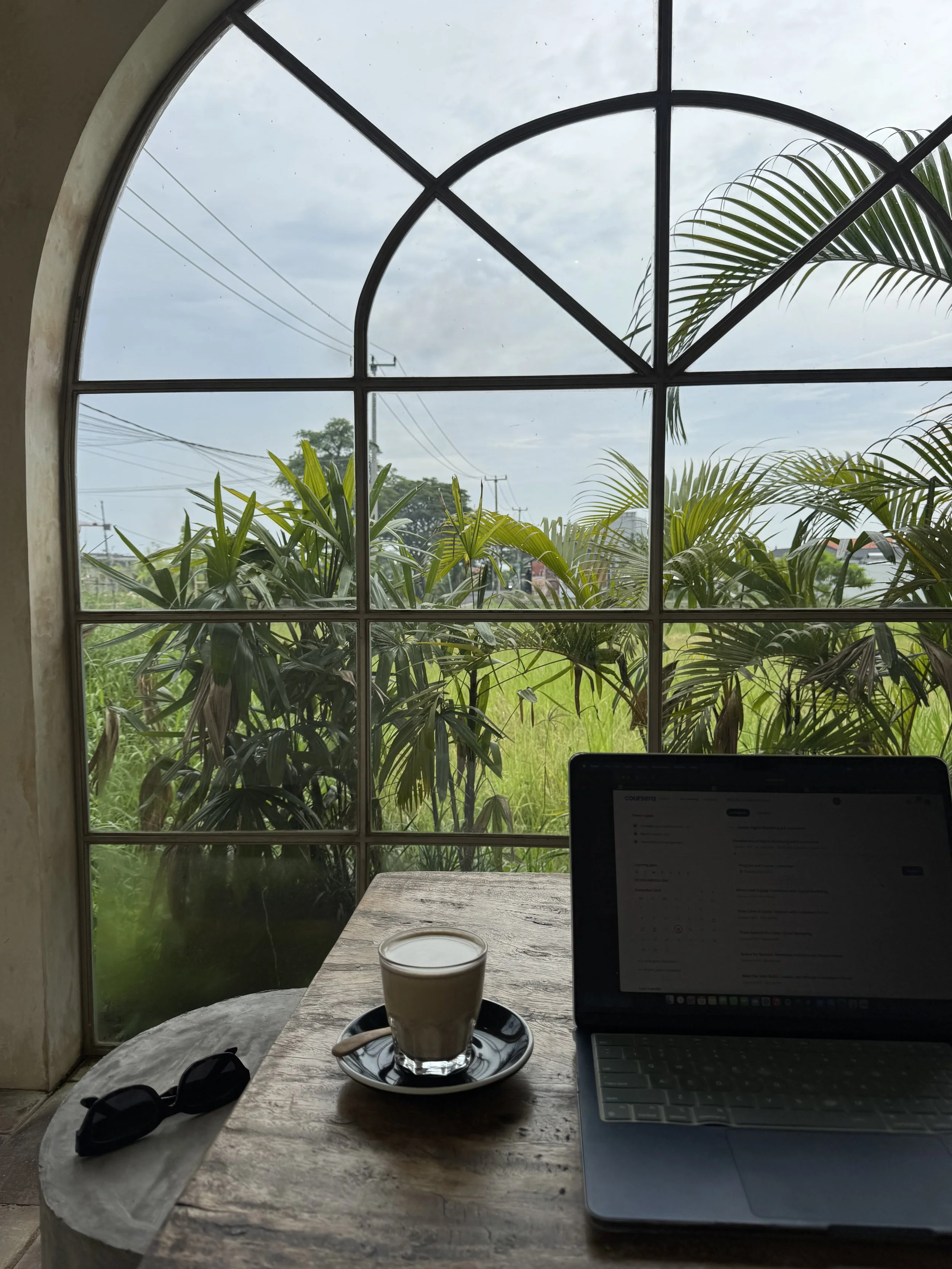 Black MacBook on a wooden table overlooking greenery and rice field, with a coffee in a transparent glass to the left and black sunglasses laid next to the coffee