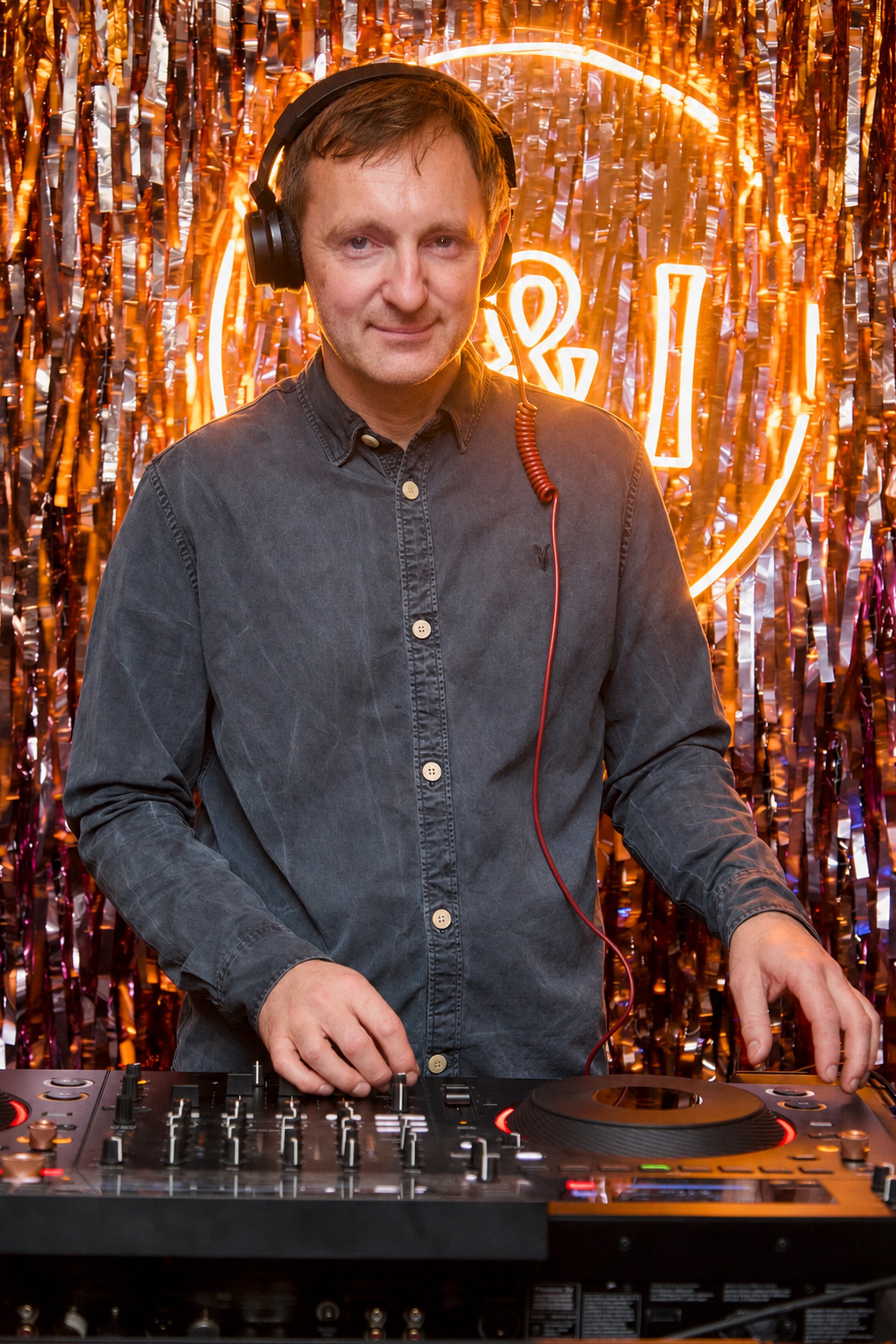 A man DJing at a wedding with a copper and orange tinsel backdrop and neon orange lights forming a shape behind him.