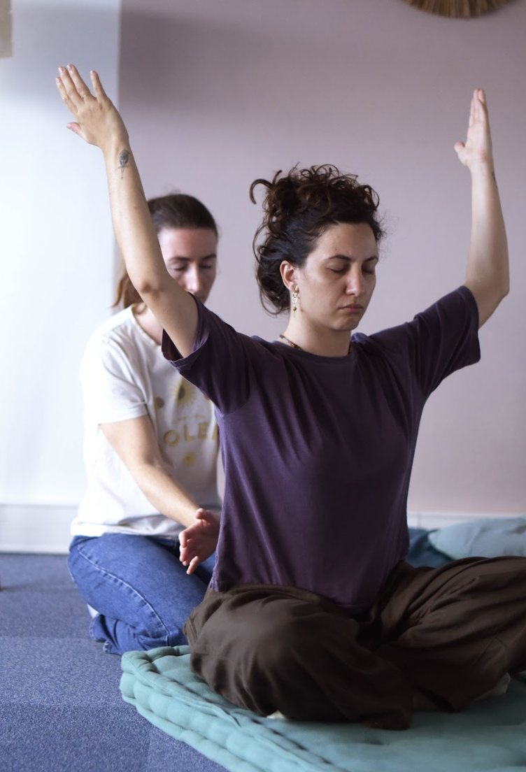 Une femme médite assise en position du lotus avec les yeux fermés, bras levés, pendant qu'une autre femme est derrière elle, en arrière-plan, dans une pièce calme.