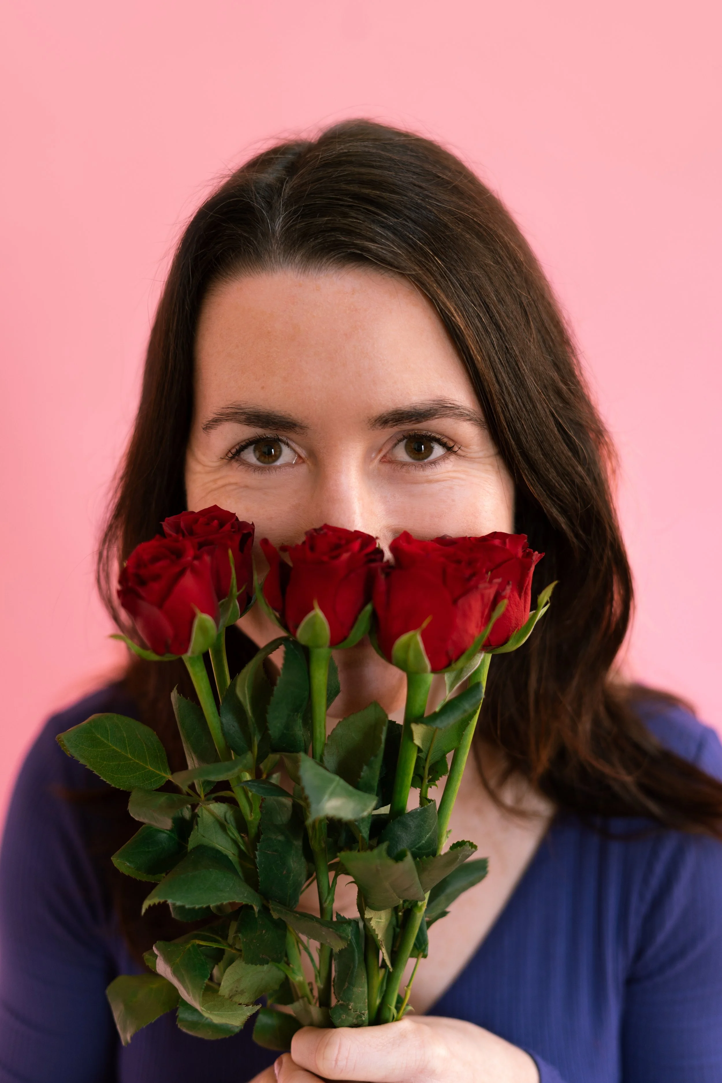 Femme tenant un bouquet de roses rouges devant son visage, yeux visibles, fond rose.