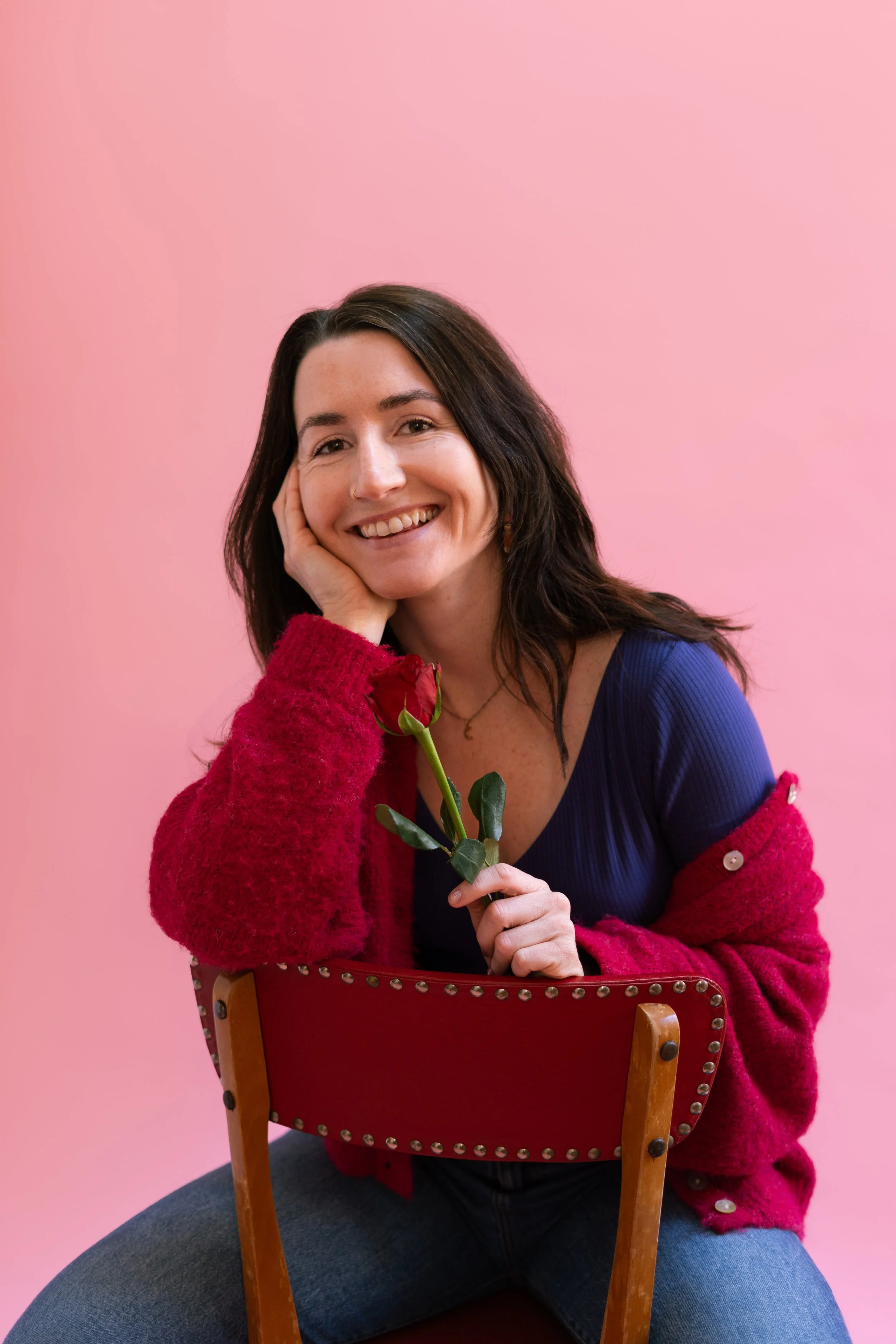 Une femme souriante avec des cheveux bruns, portant un pull rouge et un vêtement bleu, tenant une rose rouge, assise sur une chaise contre un fond rose.
