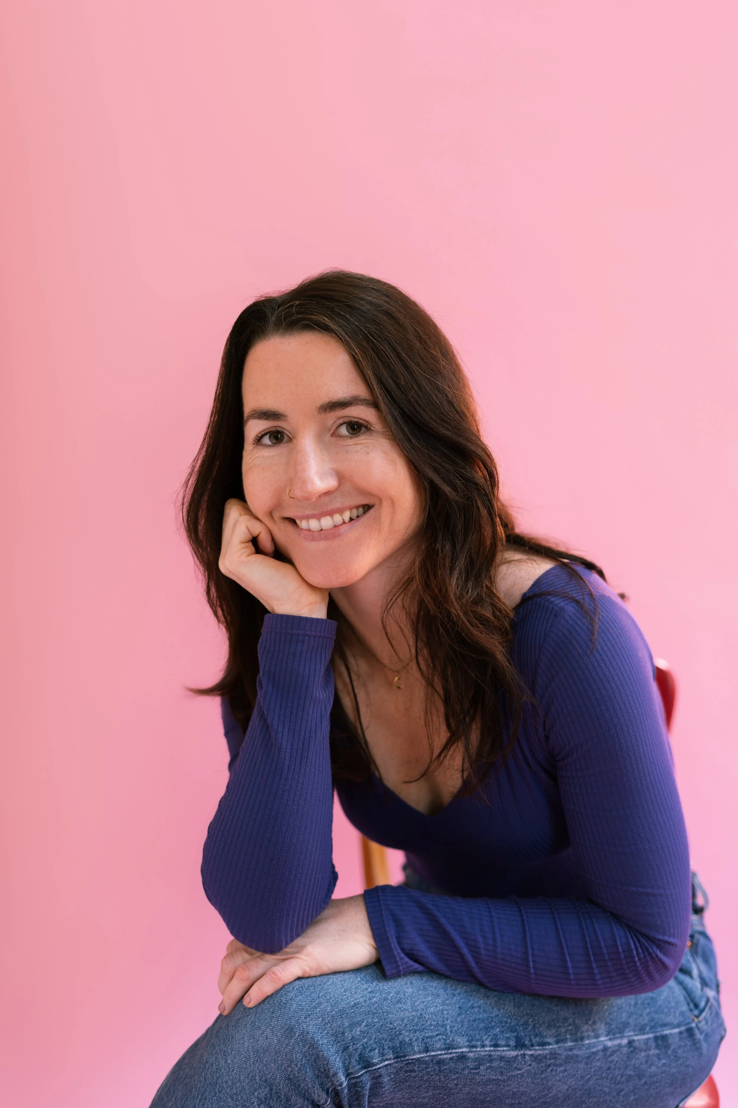 Une femme souriante avec les cheveux bruns longs, portant un pull violet et assise sur une chaise, contre un fond rose