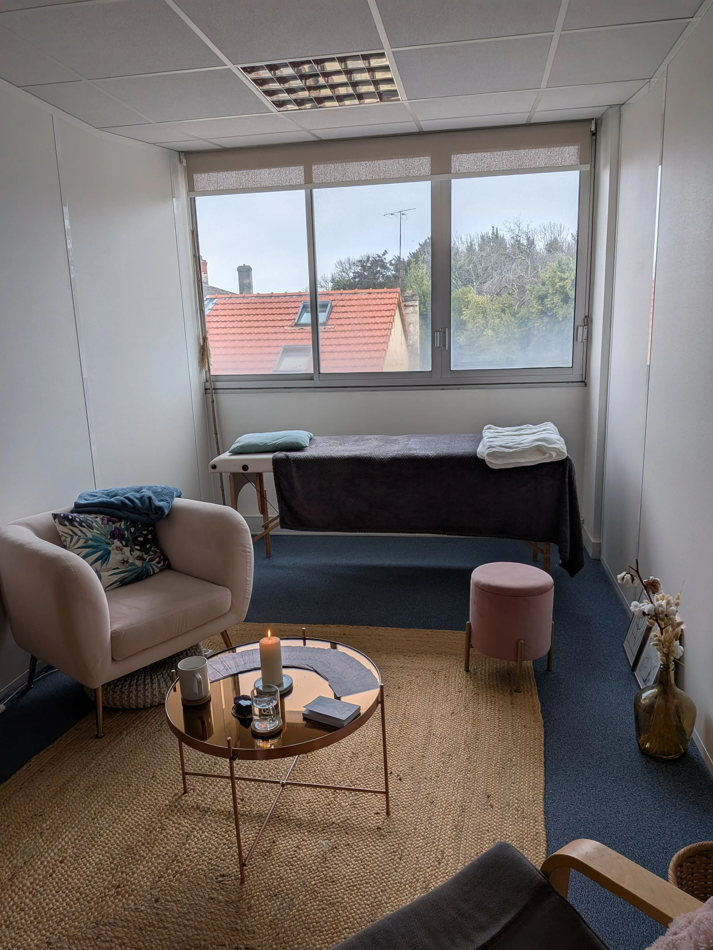 Salle de relaxation avec un canapé, une table ronde, une bougie allumée, un fauteuil, une fenêtre avec vue sur les toits et la nature extérieure, un lit de massage, une petite banquette rose, et un vase avec des fleurs