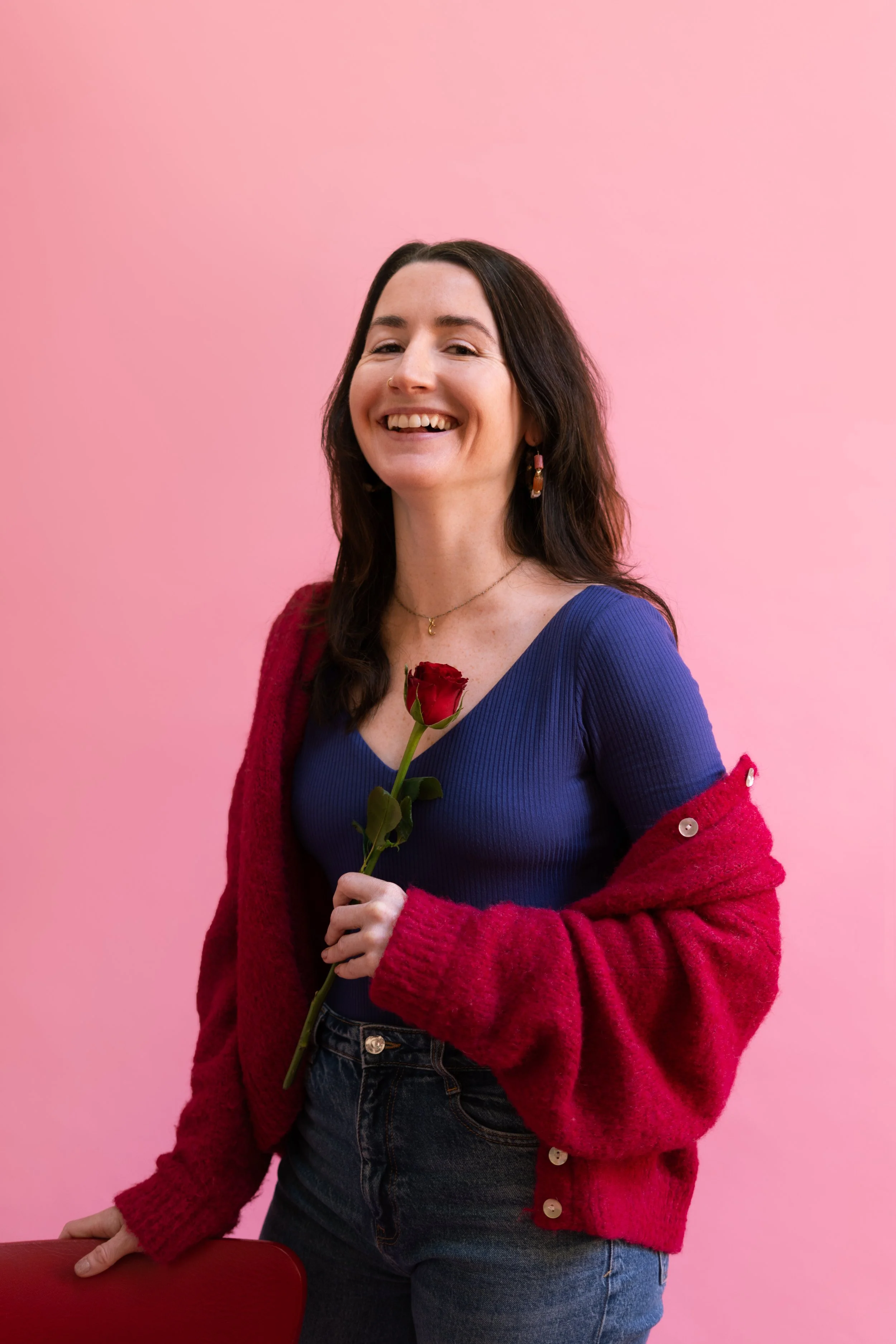 Une femme souriante tenant une rose rouge devant un fond rose.
