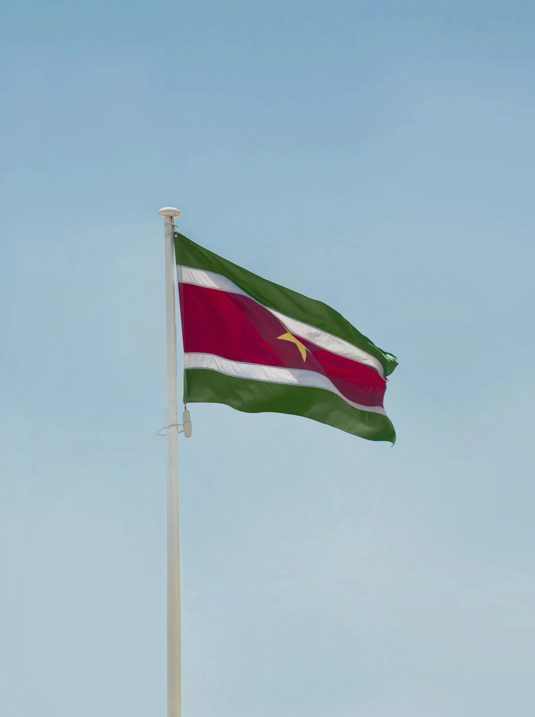 een vlag met groene, witte, rode en gele kleuren wappert in de wind, bevestigd aan een witte vlaggenstok tegen een blauwe lucht. A&R Chicken and More is een lokaal Surinaams bedrijf dat gespecialiseerd is in marinade en kipproducten.