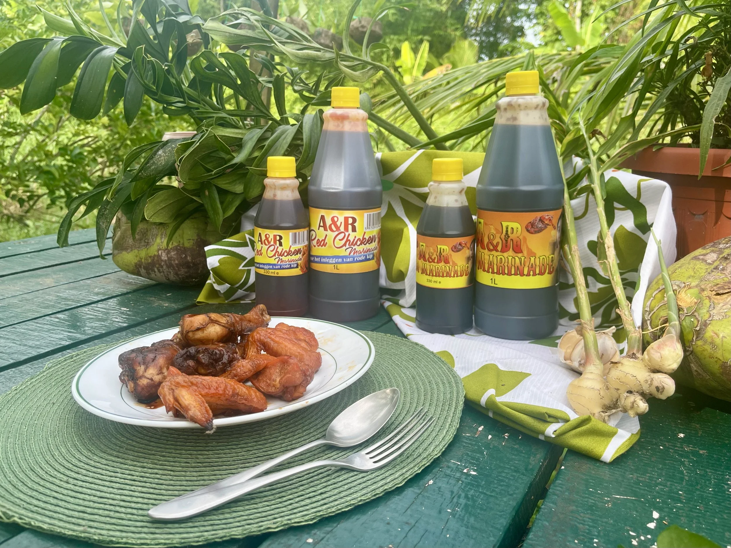 Gekookte kippenvleugels en drumsticks op een bord, omringd door marinadeflessen van A&R Chicken and More, groene planten, gember en een groene tafel. A&R Chicken and More exporteert naar Nederland en verkoopt lokaal in Suriname.
