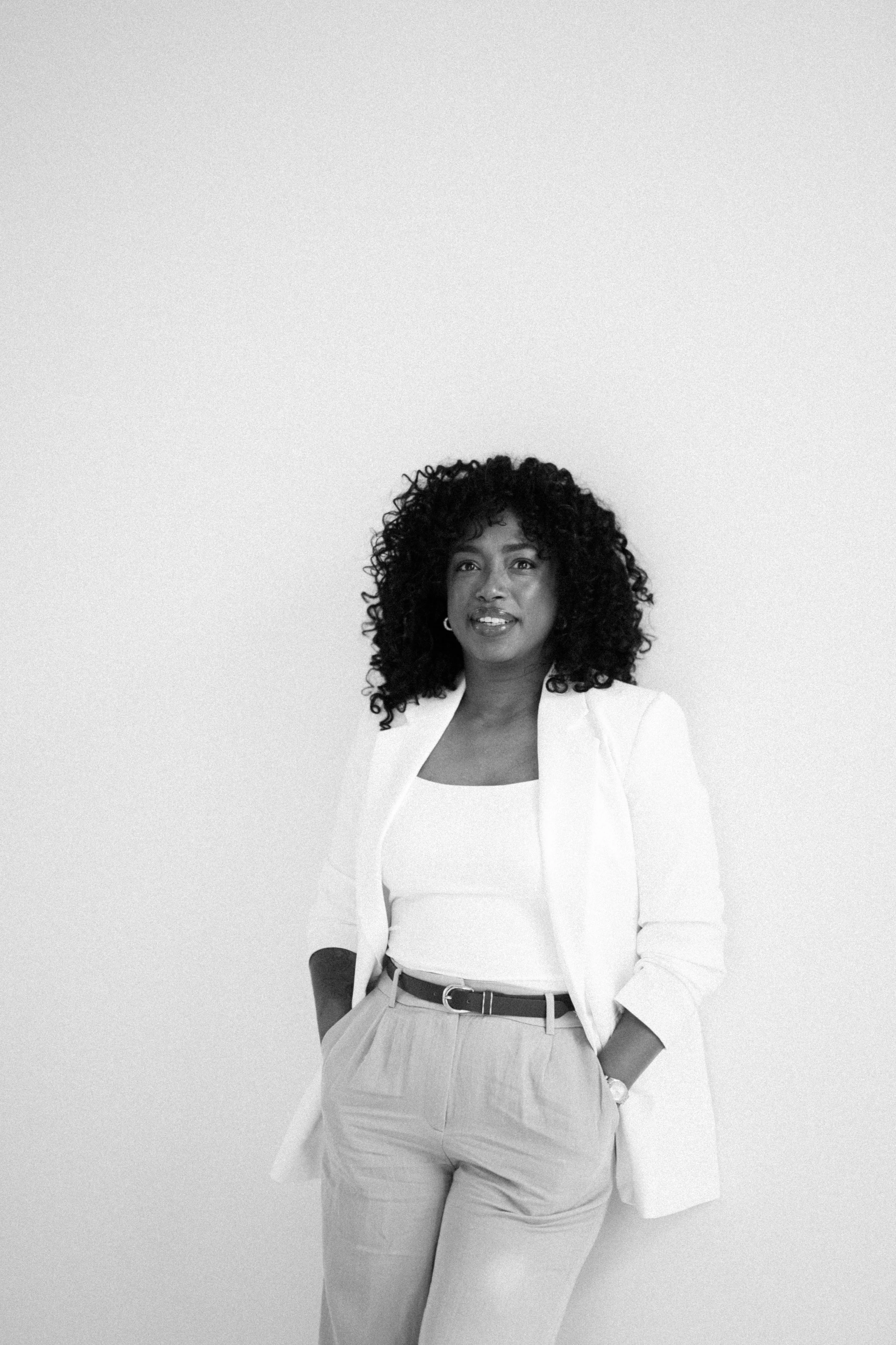 Zwarte vrouw met gekruld haar gekleed in een witte blazer en pantalon, poseert voor een witte achtergrond.