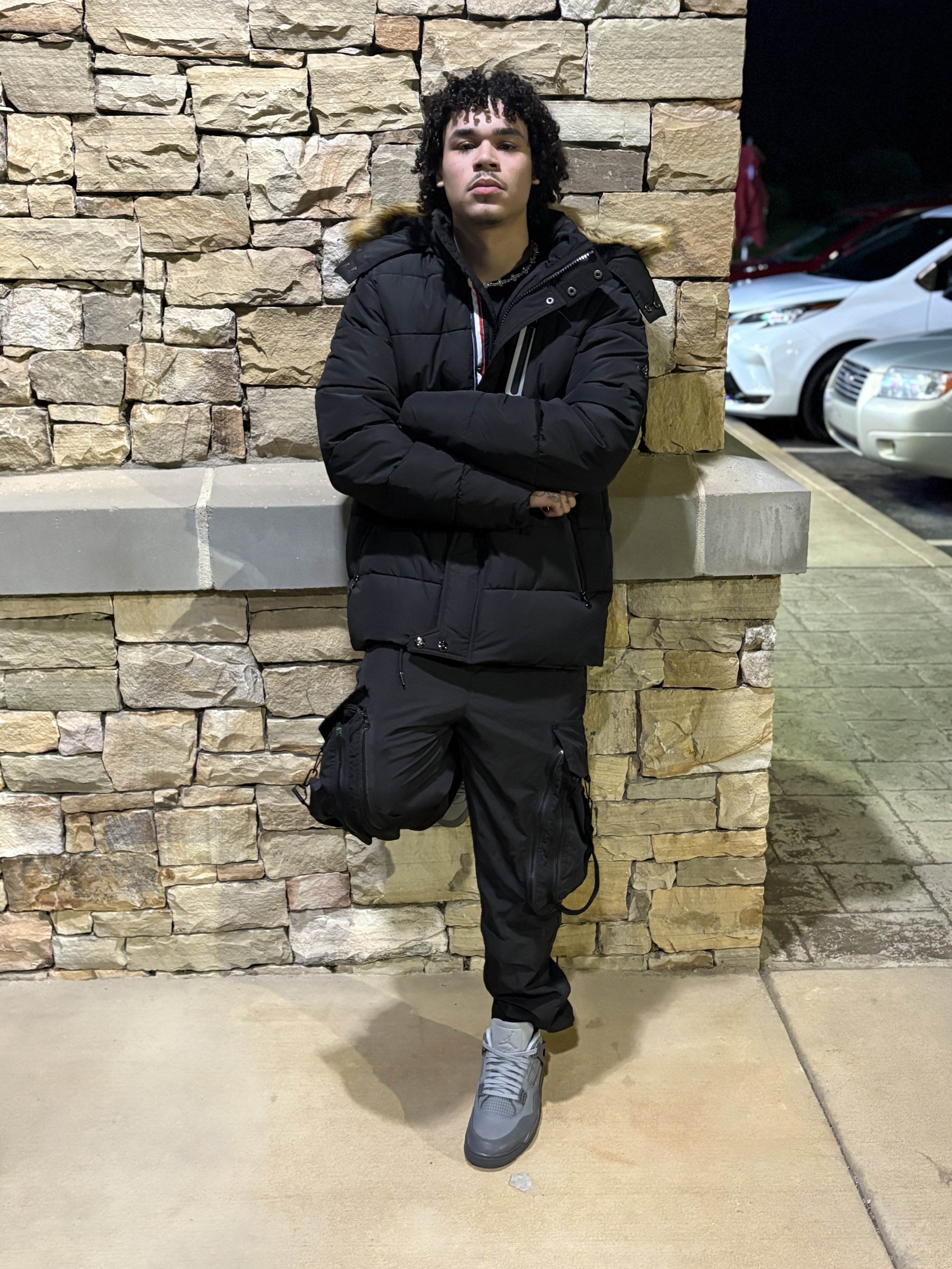 A young man with curly hair, dressed in a black puffy jacket, black cargo pants, and gray sneakers, leaning against a stone wall with arms crossed.