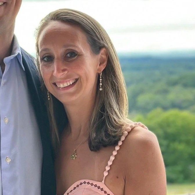 A smiling woman with shoulder-length brown hair, wearing drop earrings, a necklace, and a pink dress with spaghetti straps, standing outdoors with a blurred green landscape in the background.