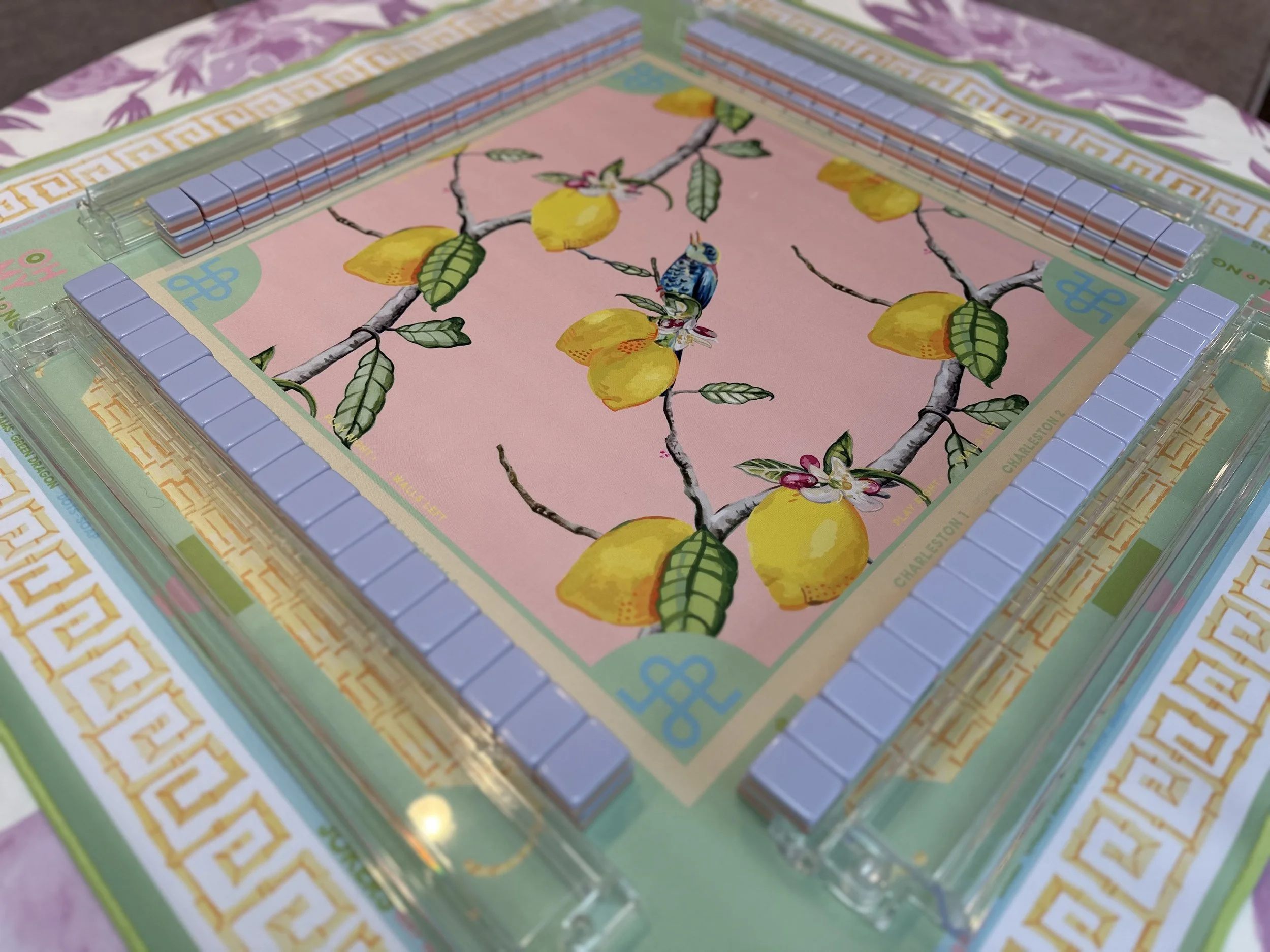 A Mah jong game set on a table with a decorative fruit and bird illustration on the playing surface, surrounded by mah jong tiles.