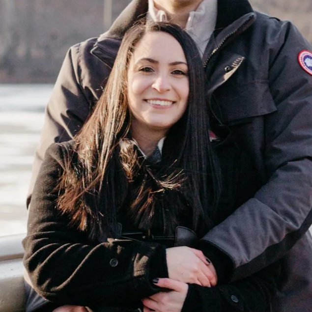 A smiling woman with long dark hair being hugged by a person wearing a dark jacket with a Canada Goose logo, outdoors near water.