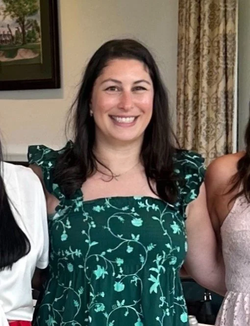 Smiling woman with long dark hair wearing a teal dress with white floral patterns, standing indoors with beige curtains and a framed picture in the background.
