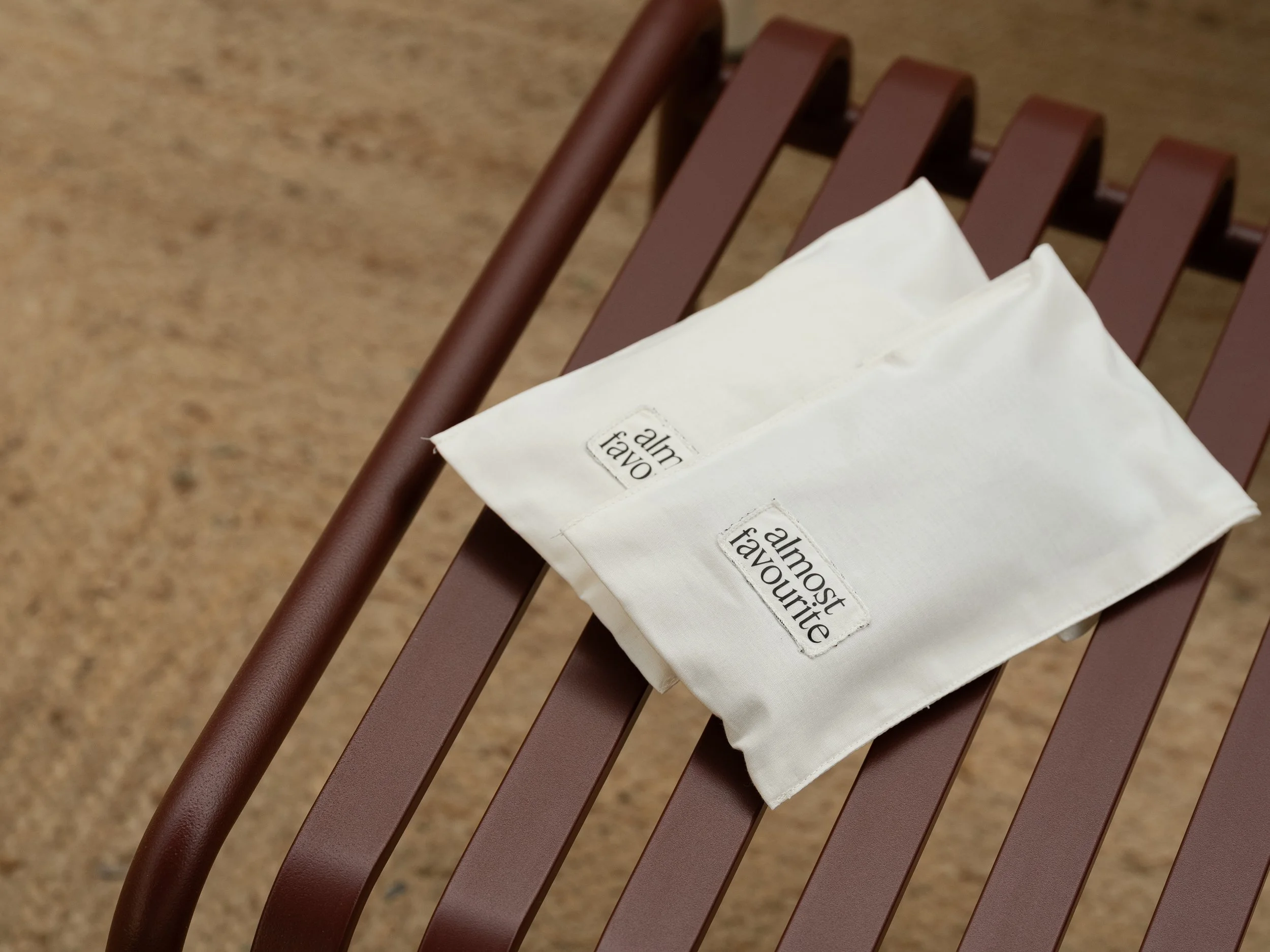 Two white cloth bags with black embroidered text that says 'almost favourite,' placed on a brown metal bench with slats.