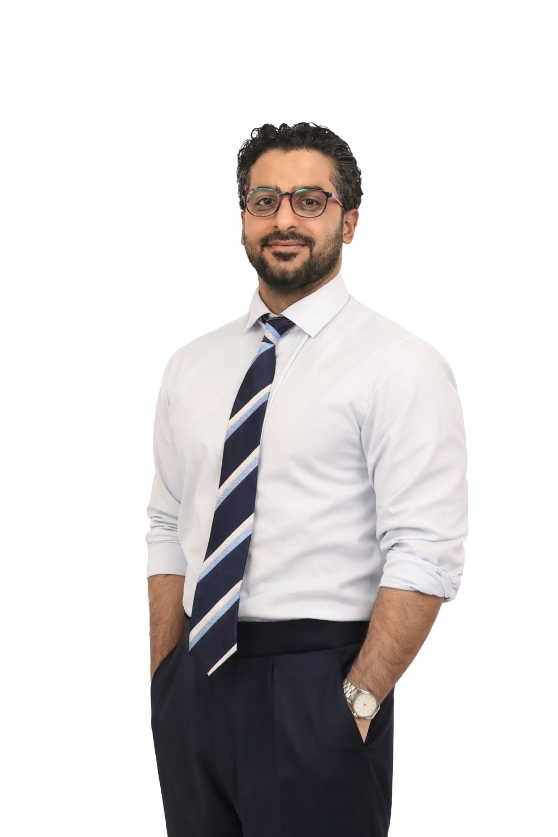 Moh'd Ghayeb wearing a white shirt, a striped tie, and dark pants, standing with his hands in his pockets against a solid background.