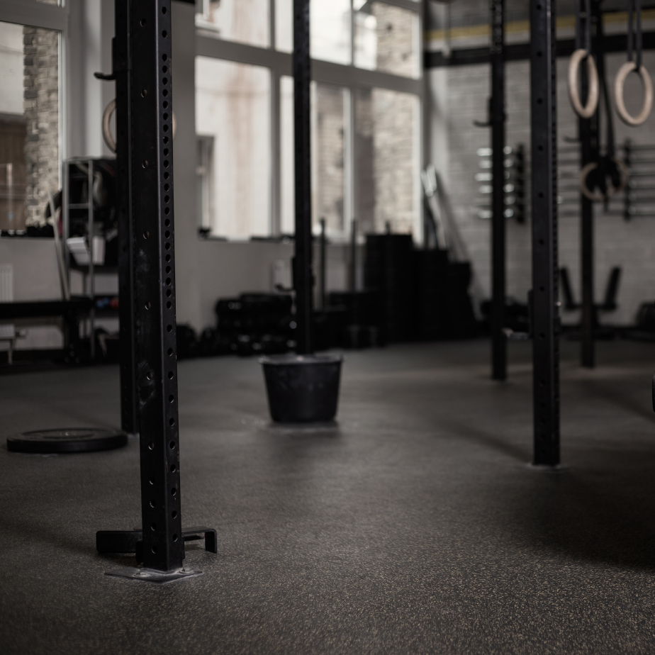 Empty gym with black metal squat rack, black rubber-coated weights, black gym bucket, and gymnastic rings hanging in background.
