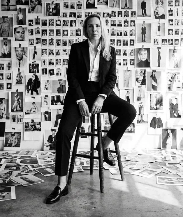 A woman in a black suit and white shirt sitting on a wooden stool in front of a wall covered with black-and-white photographs, with scattered photos on the floor.